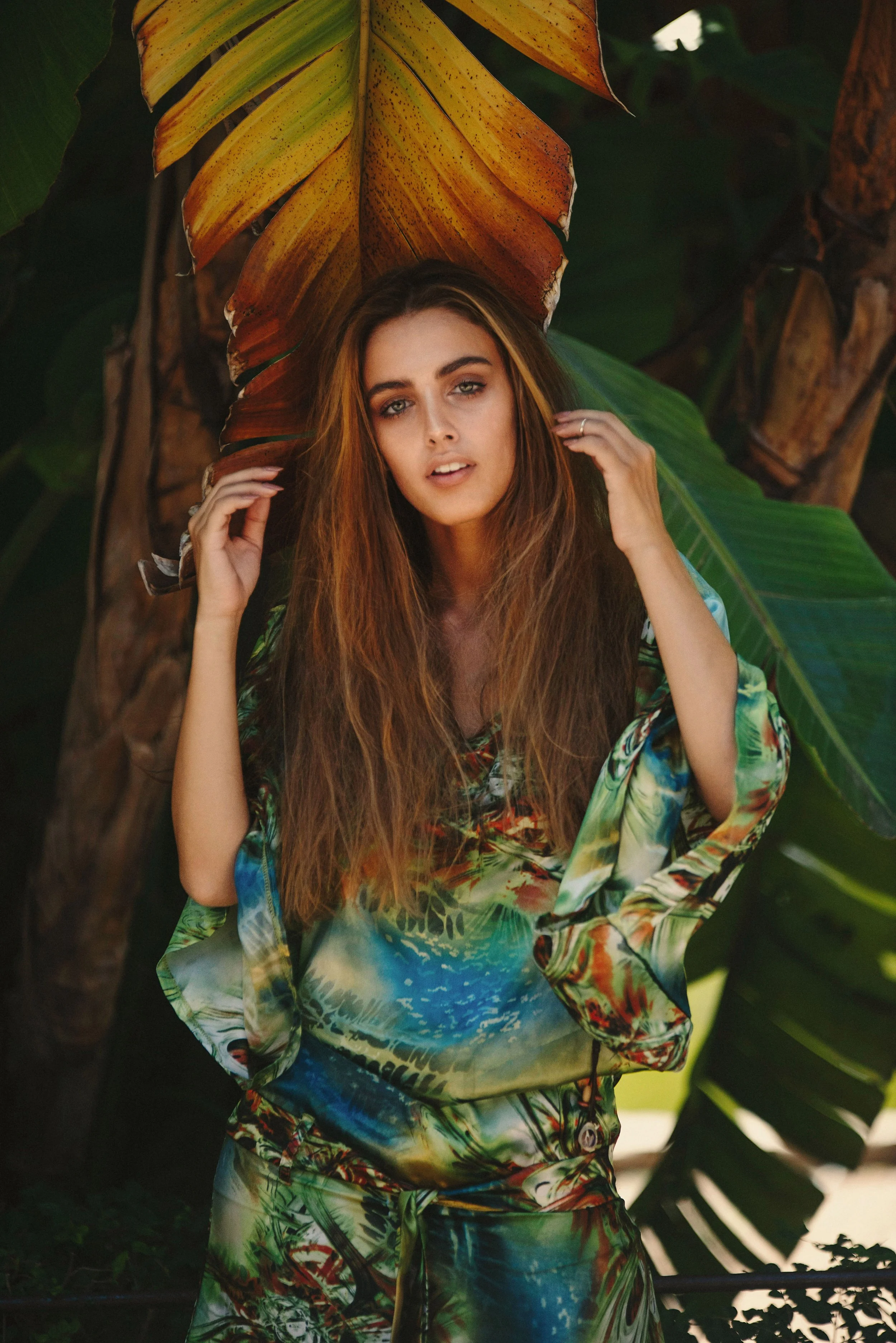 A young woman with long brown hair, wearing a colorful, patterned outfit, standing in front of large green and orange tropical leaves, posing outdoors.