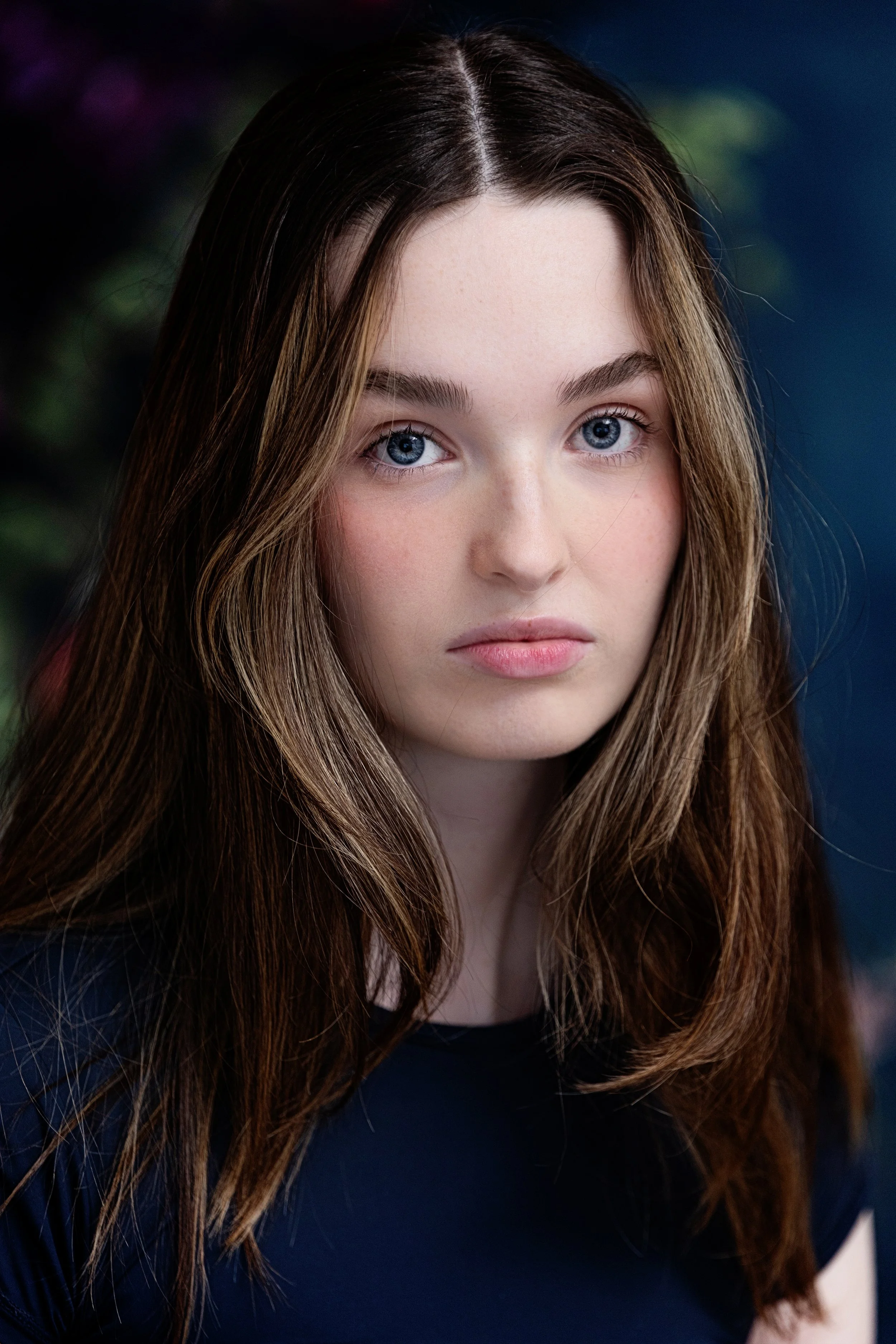 A young woman with long, wavy brown hair and blue eyes looking at the camera, wearing a dark top, with dark blurred background.