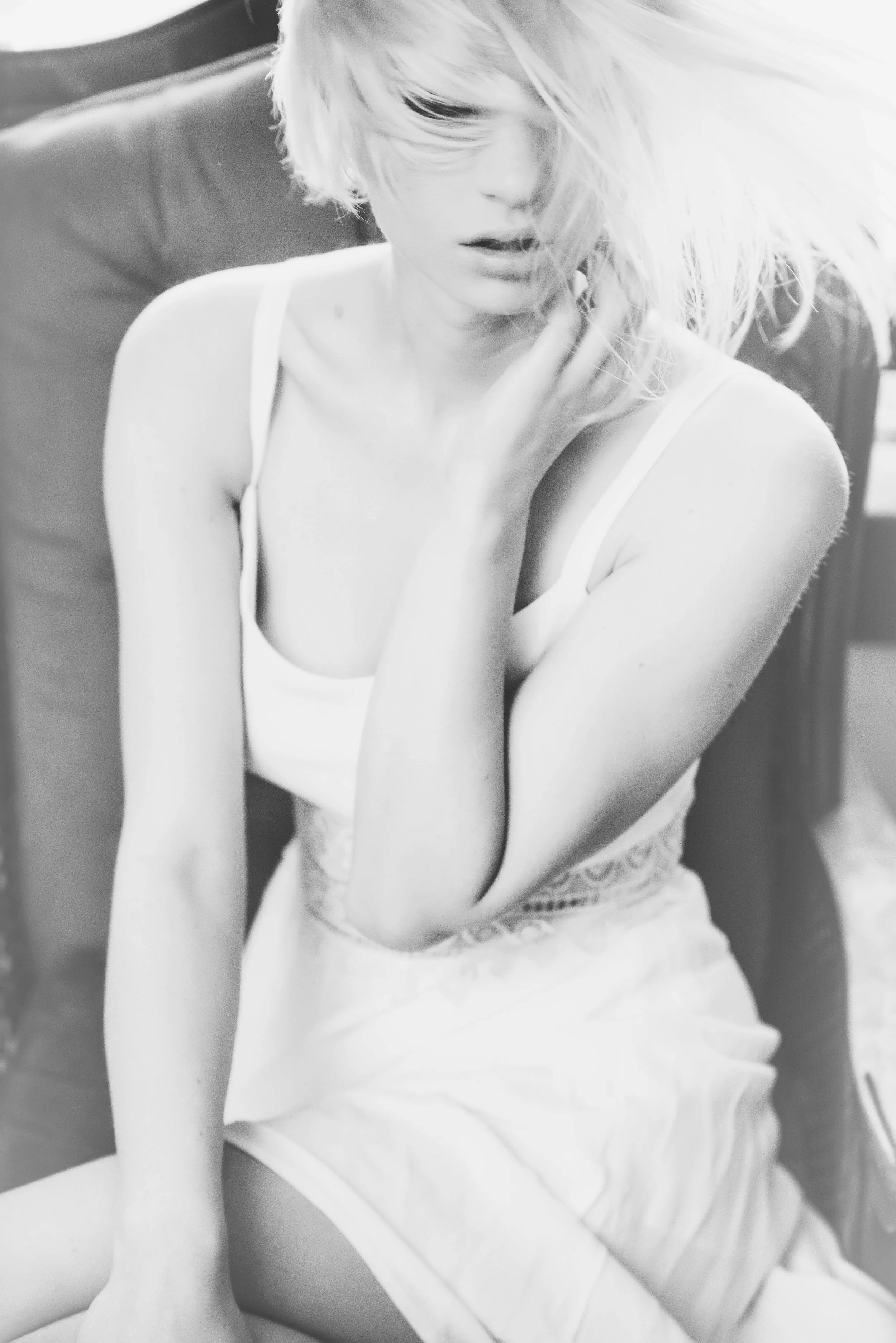 A woman with blonde hair covering part of her face, wearing a white tank top and shorts, sitting on a bed with a gray blanket. She is looking down and slightly to the side, with her hand touching her neck.