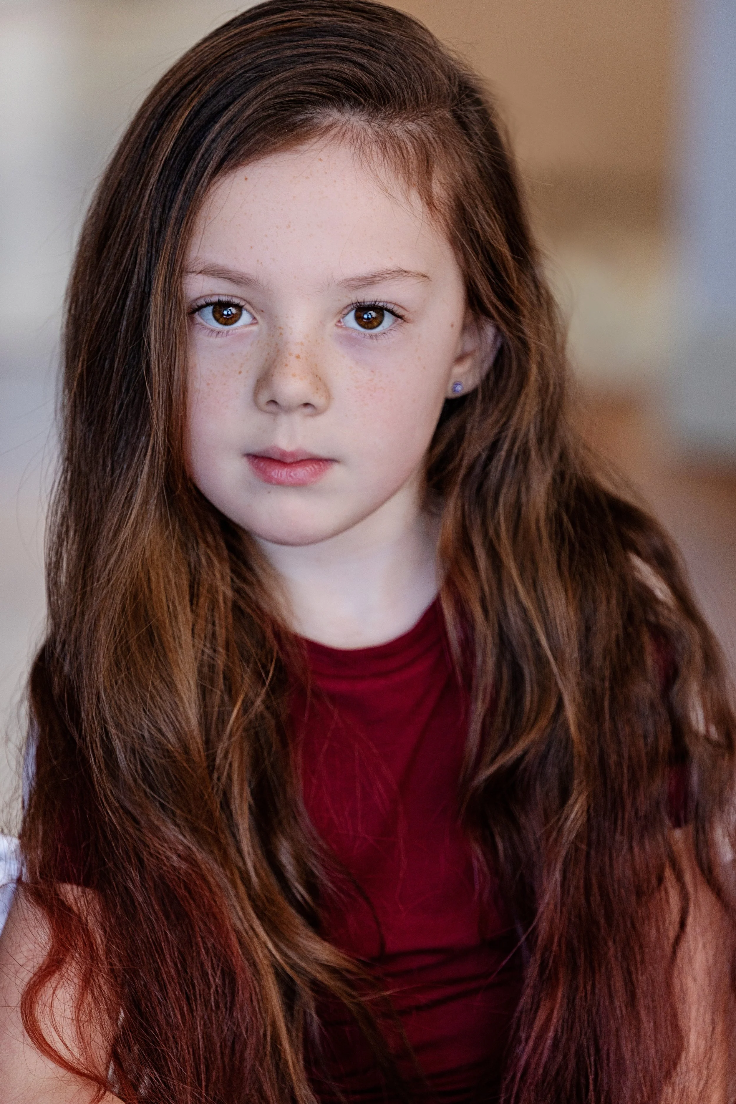 Close-up of a young girl with long, wavy auburn hair, brown eyes, and freckles, wearing a burgundy top and small earrings.
