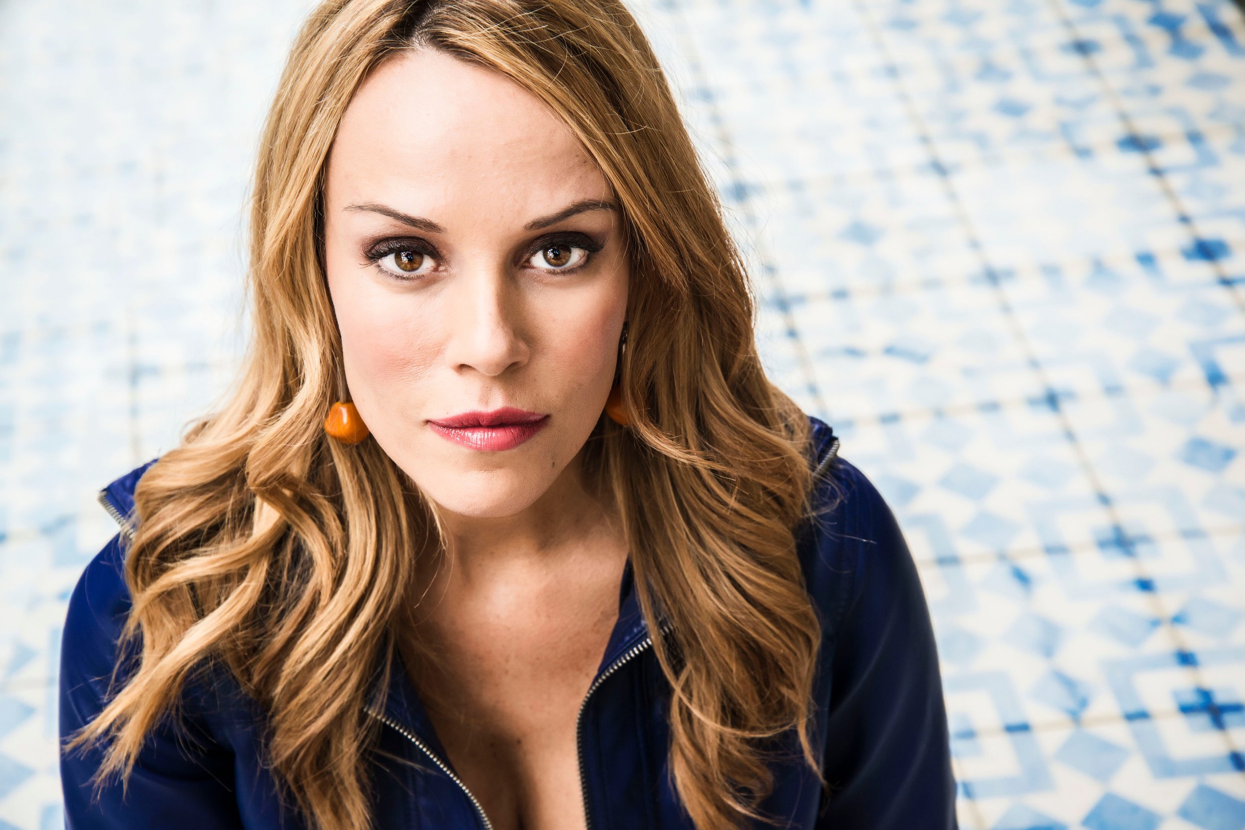 Close-up of a woman with long wavy red hair and brown eyes, wearing a dark blue zip-up jacket and orange earrings, standing in front of a tiled wall.