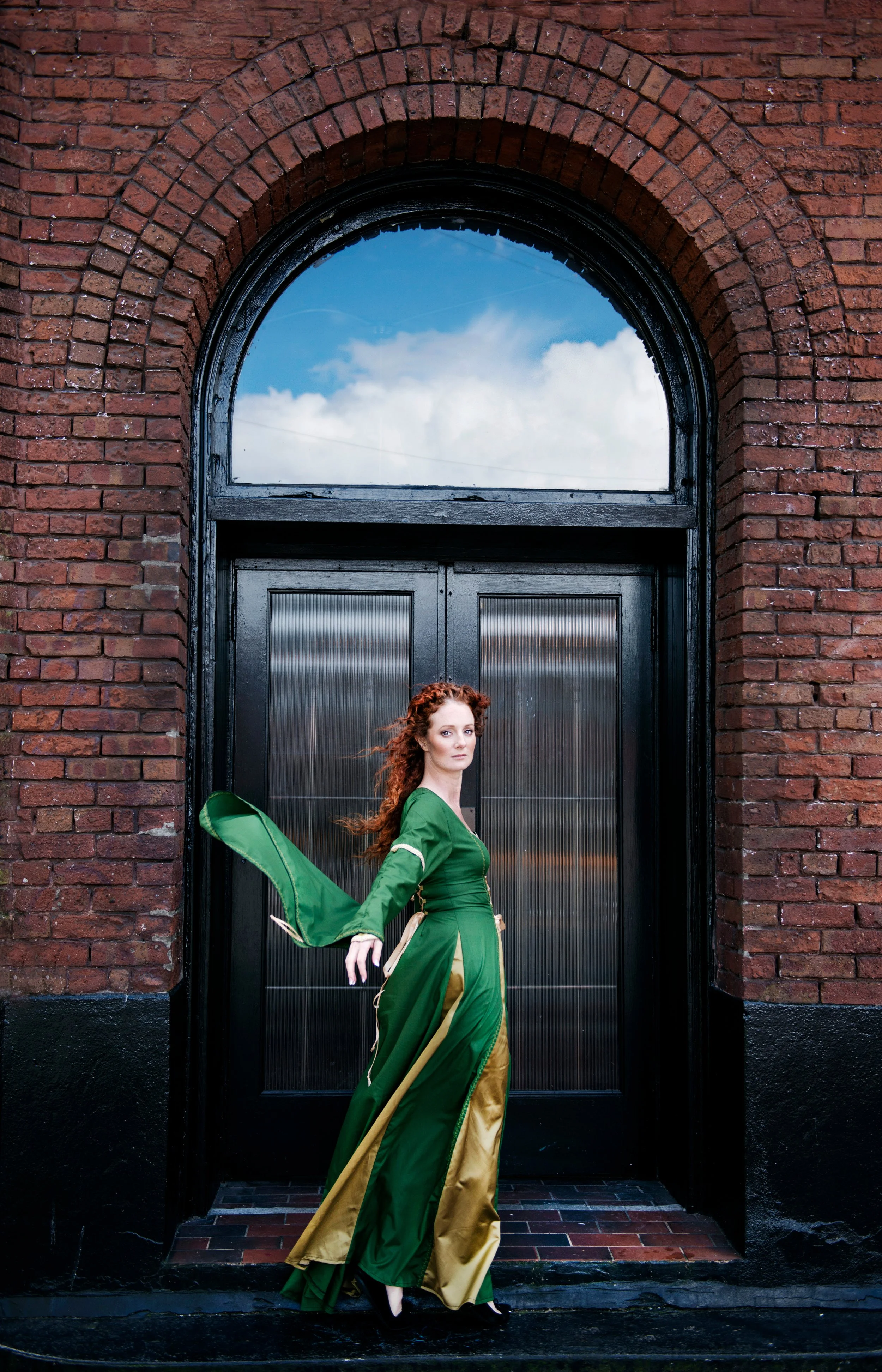 A woman with curly red hair wearing a green and gold costume with a cape, standing in front of a black door set in a red brick building, with a large arched window above the door reflecting a partly cloudy sky.