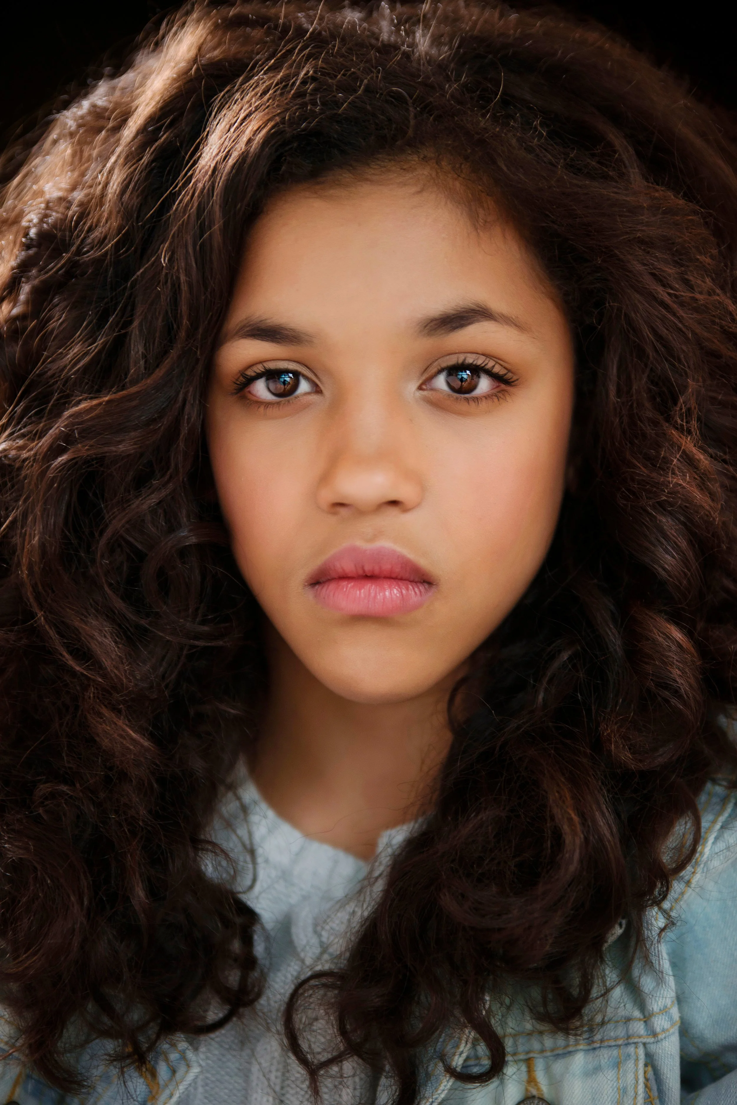 Close-up of a young woman with long, curly brown hair, brown eyes, and wearing a light denim jacket.