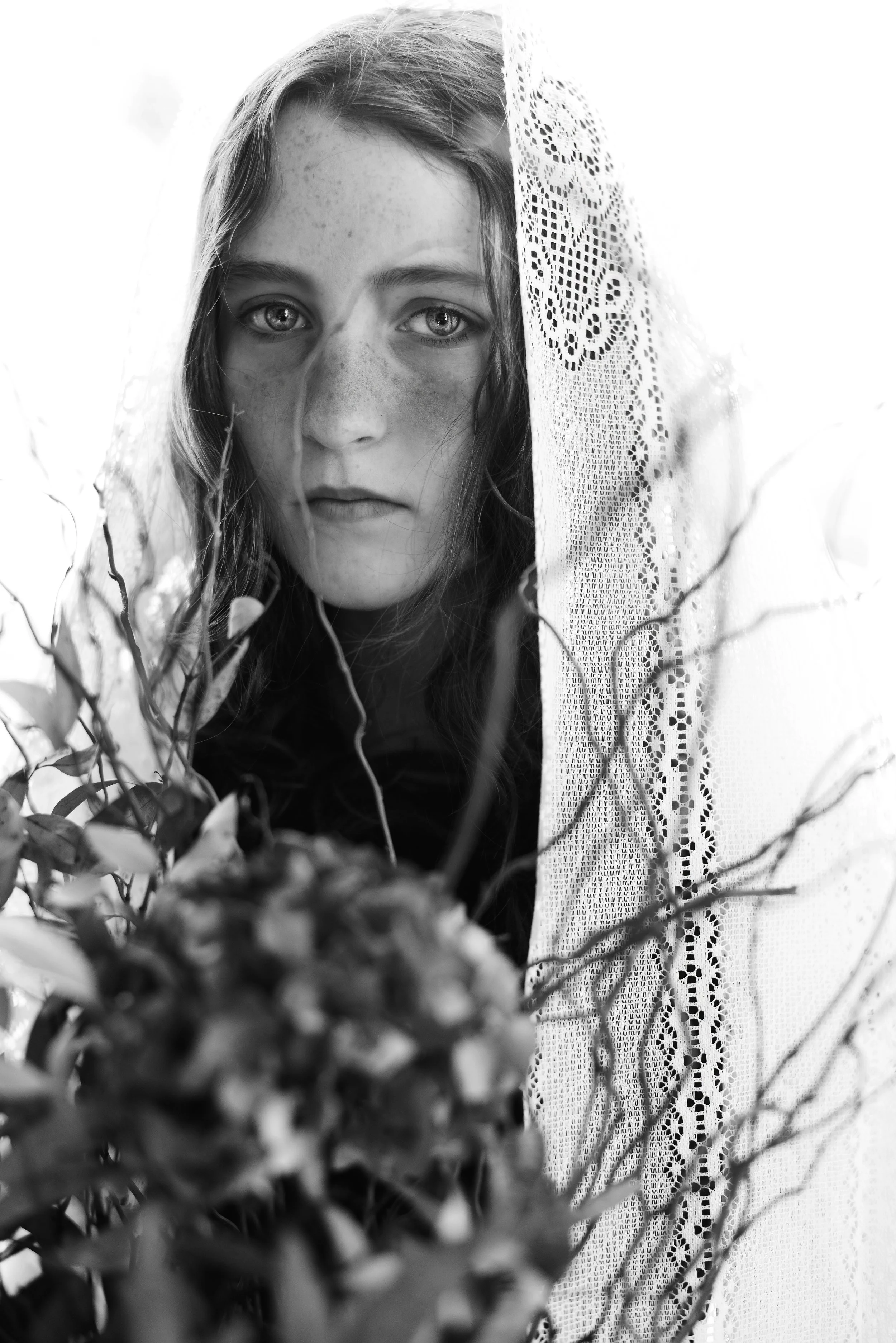 Black and white portrait of a young girl with long hair, freckles, and intense eyes, partially covered by a lace curtain and surrounded by branches and leaves.