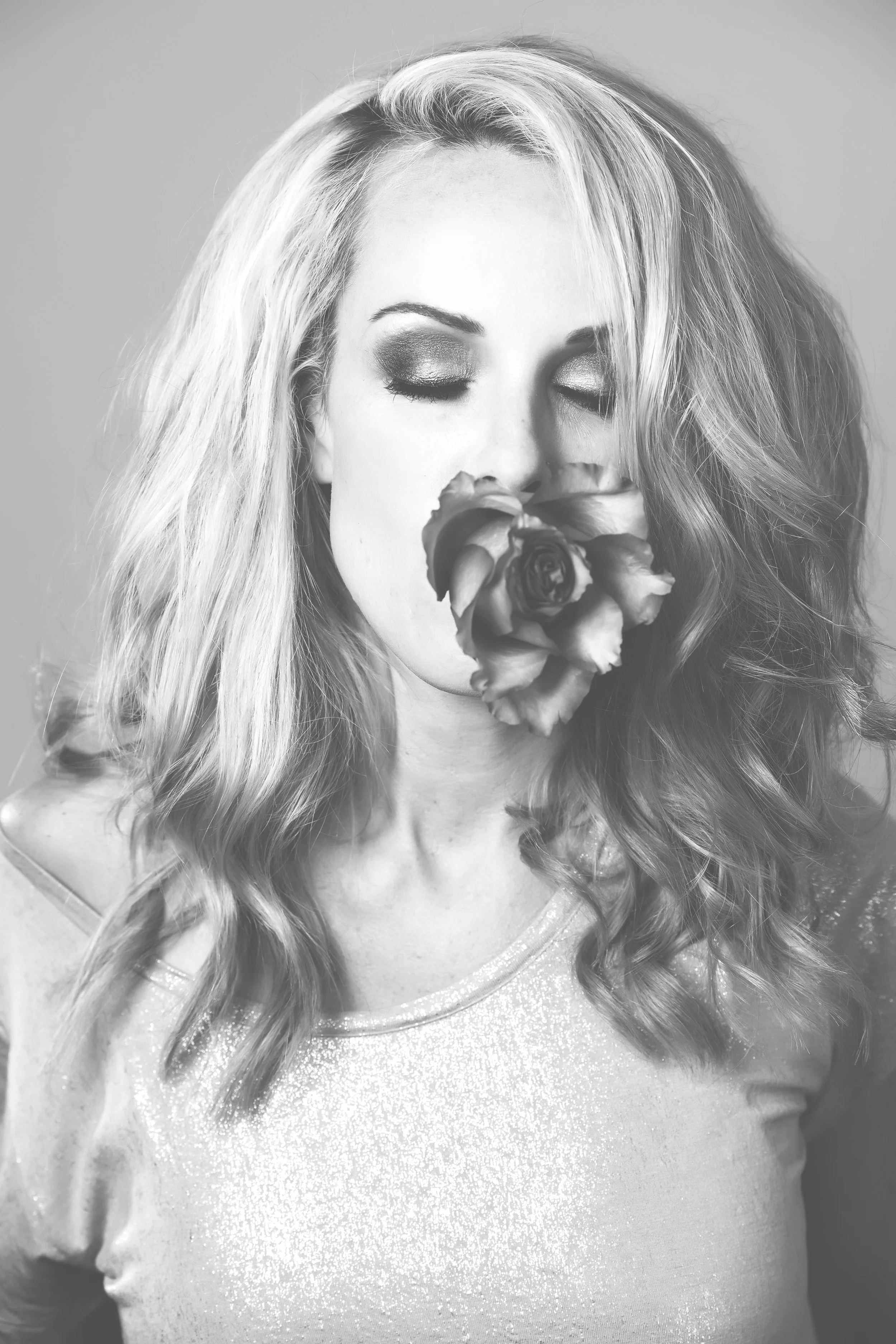 Black and white photograph of a woman with long, wavy hair, eye makeup, and a large flower held close to her face, wearing a shiny, textured top.