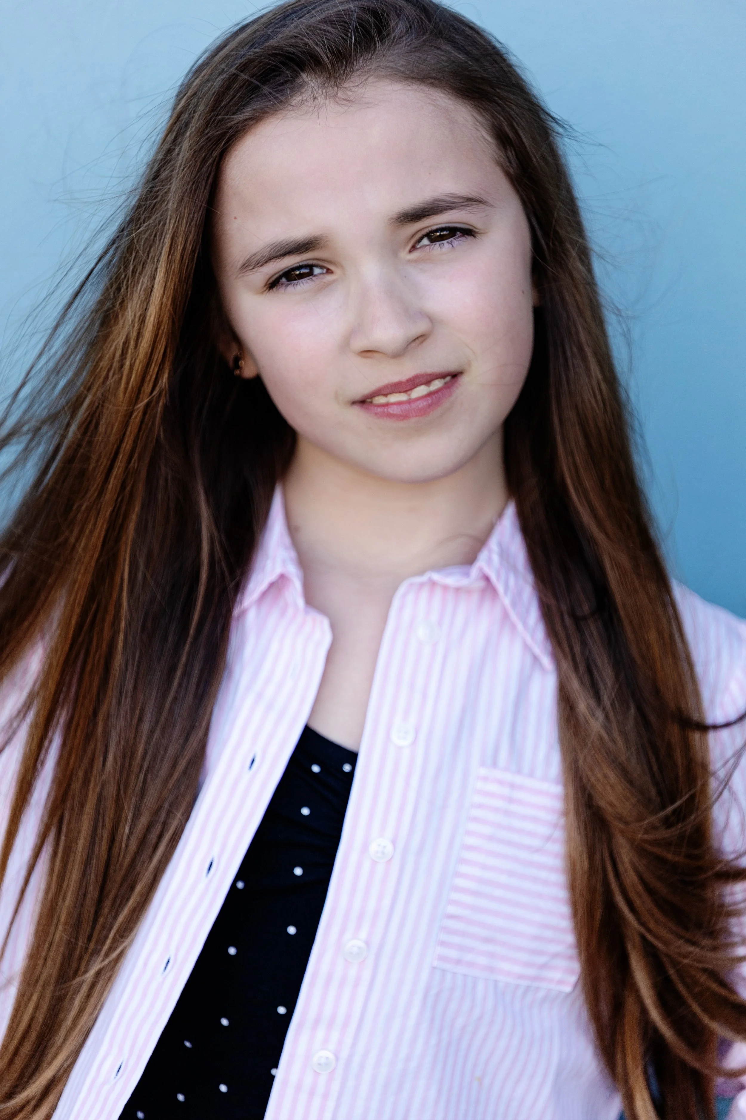 A young girl with long brown hair wearing a pink and white striped button-up shirt over a black polka dot top, standing against a light blue background.