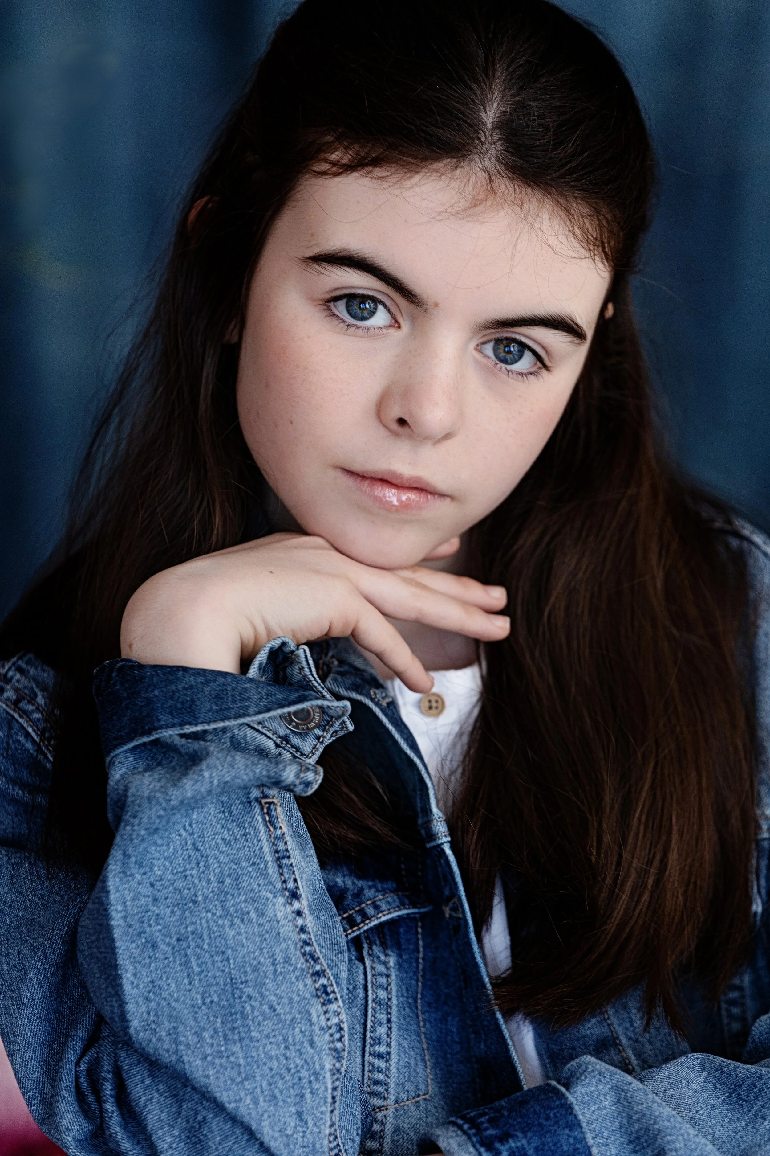 Close-up portrait of a young woman with blue eyes and long brown hair, wearing a denim jacket, resting her chin on her hand, against a dark blue background.