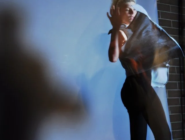 A woman standing against a wall, holding a jacket over her shoulder, looking to the side.