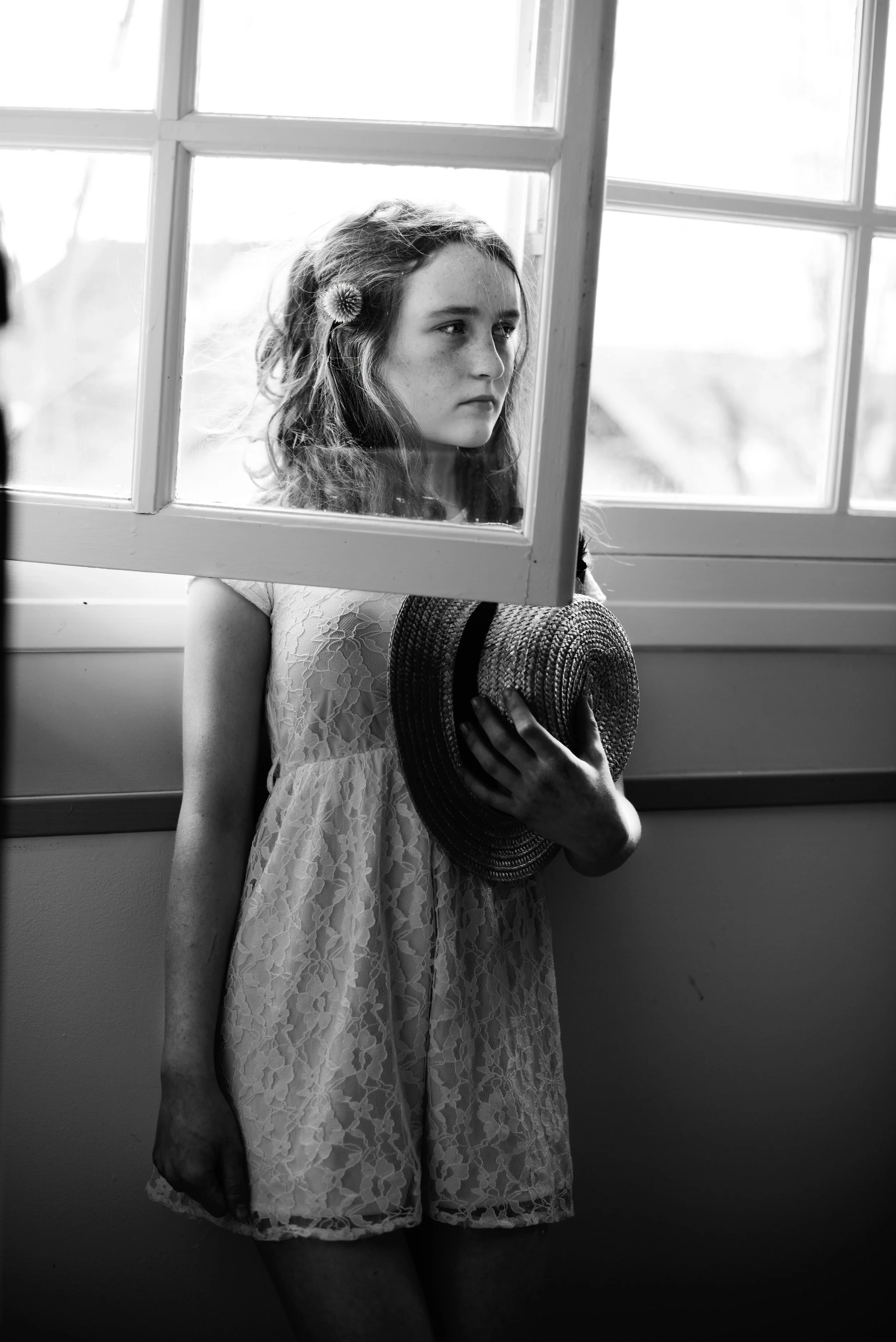 A young girl with curly hair and a flower in her hair, holding a straw hat, standing by a window in black and white.