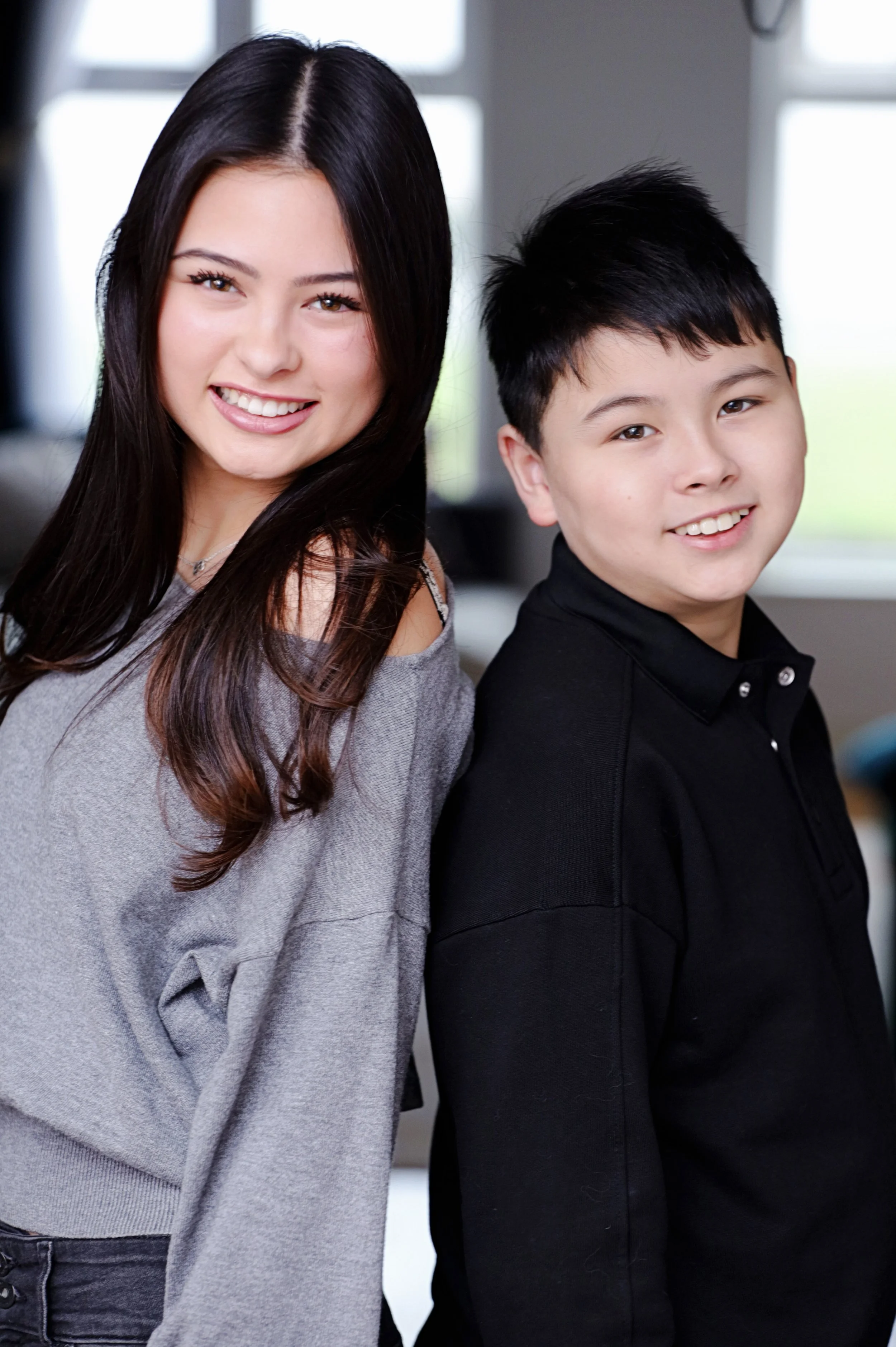A young woman with long dark hair and a young man with short dark hair, both smiling, standing indoors near windows, casual clothing.