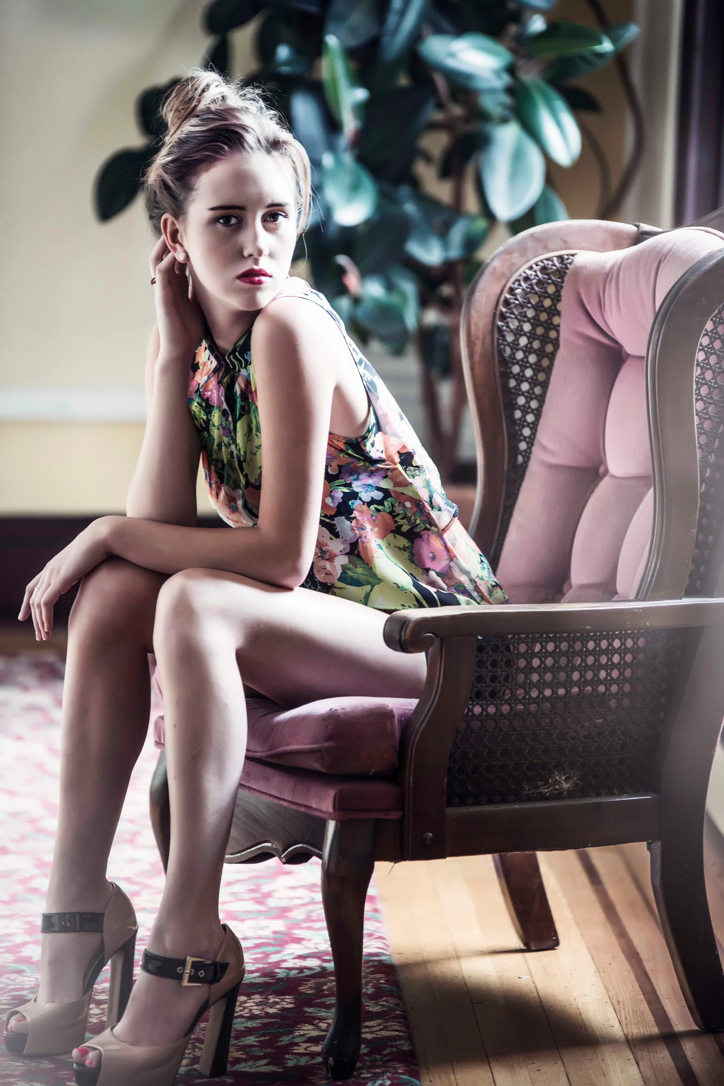 A woman sitting on a pink upholstered chair in a room with wooden floors and a patterned rug. She is wearing a floral dress, high heels, and has her hair styled up. There is a large houseplant behind her.