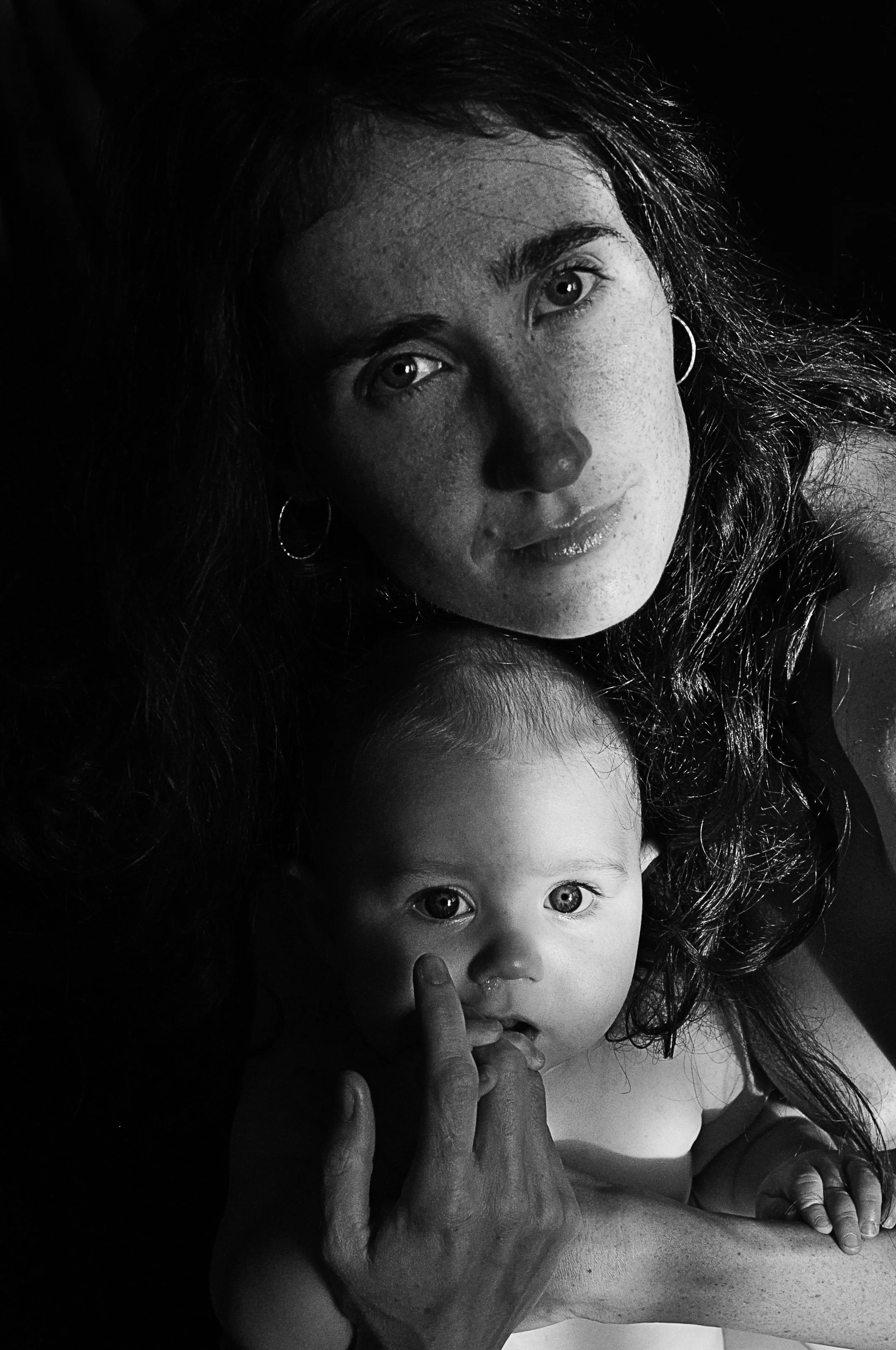 A black-and-white photo of a woman with long, curly hair and earrings, holding a young child with light-colored hair and wide eyes, both looking directly at the camera.
