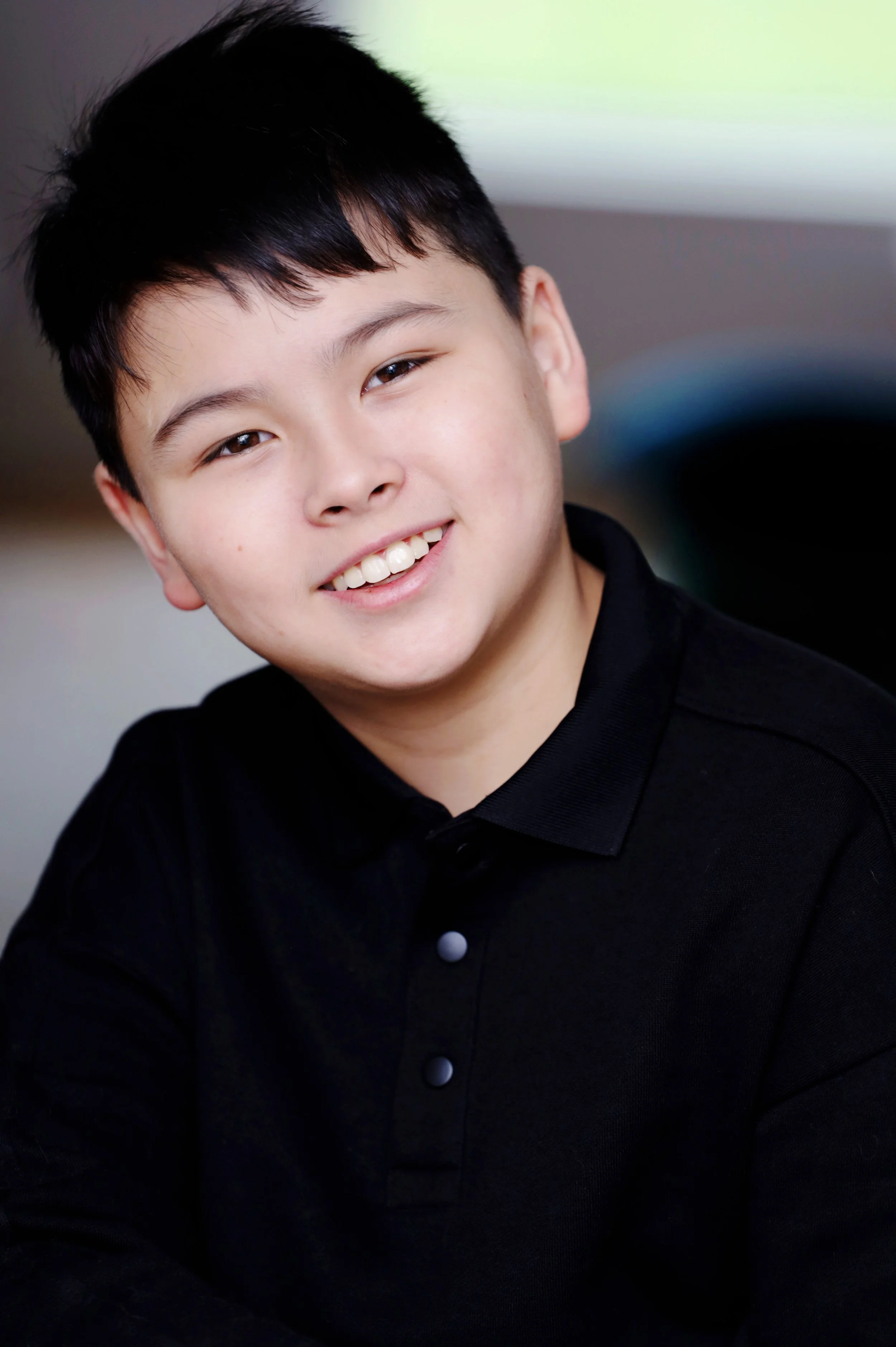 Smiling young boy with short black hair, wearing a black shirt, looking at the camera, with a blurred background.
