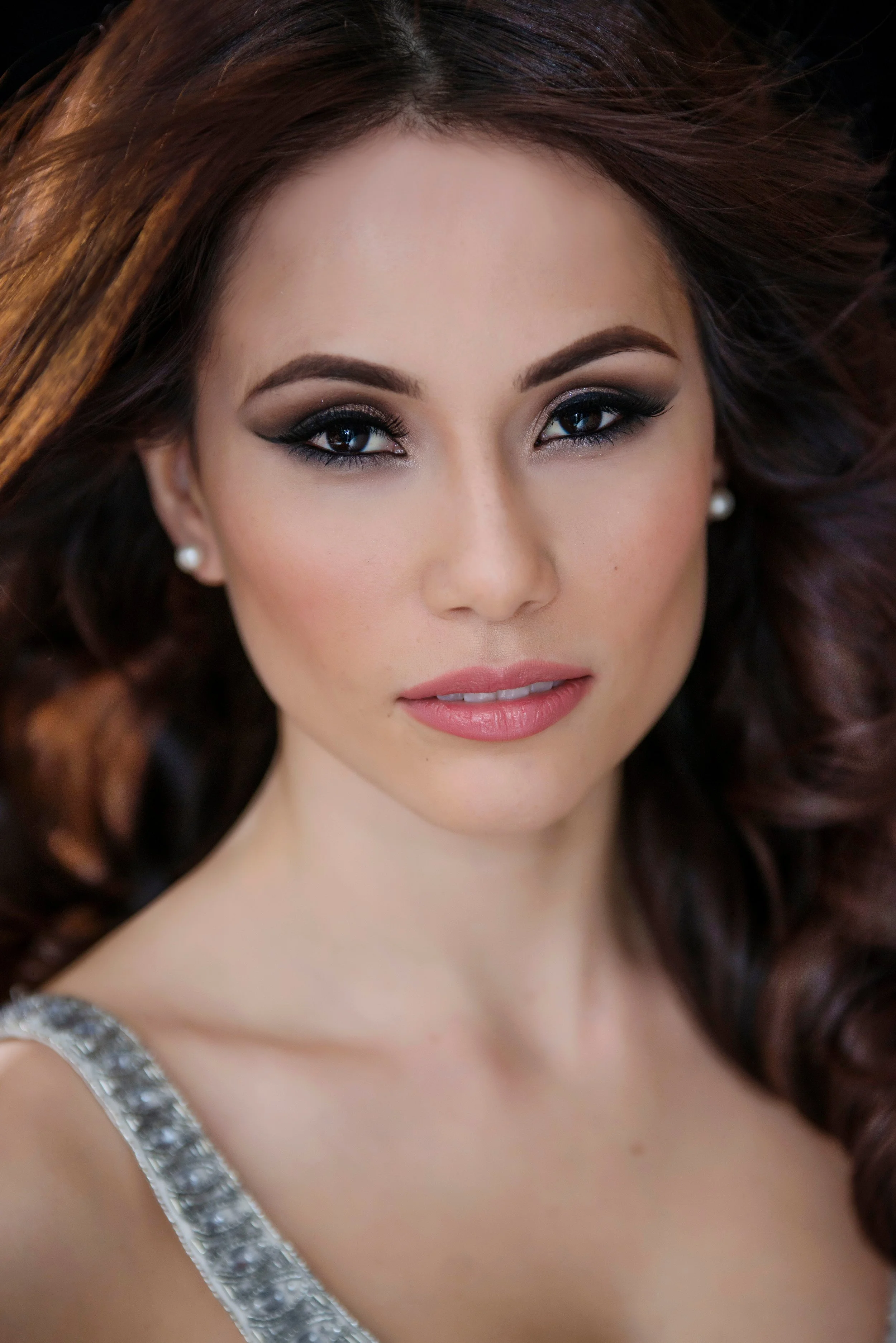 Close-up of a woman with makeup, pearl earrings, and a silver dress, looking directly at the camera with her brown hair styled in loose waves.