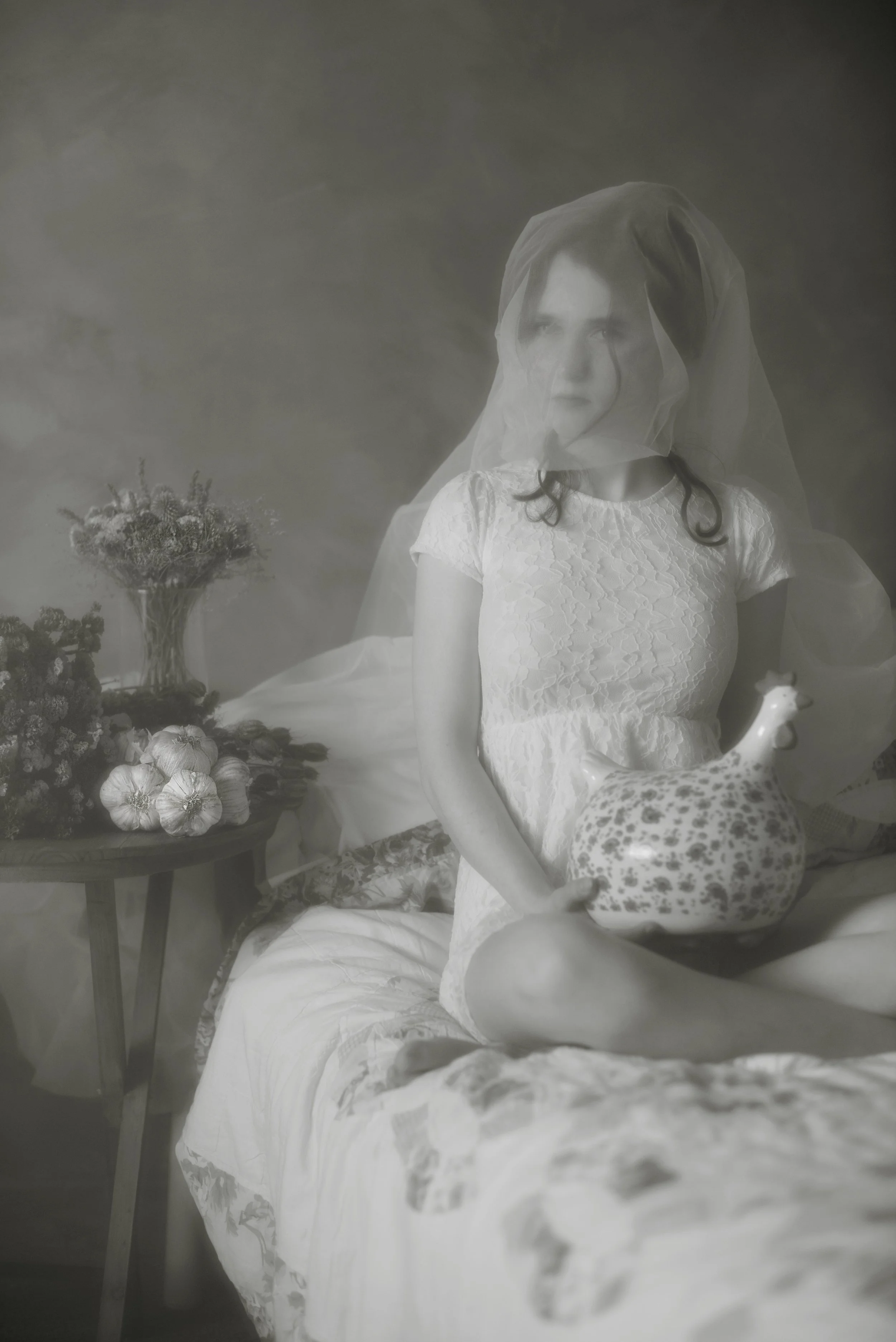 Black and white photo of a woman with dark hair covered by a sheer veil, wearing a white lace dress, sitting beside a small table with a basket of flowers and vegetables.