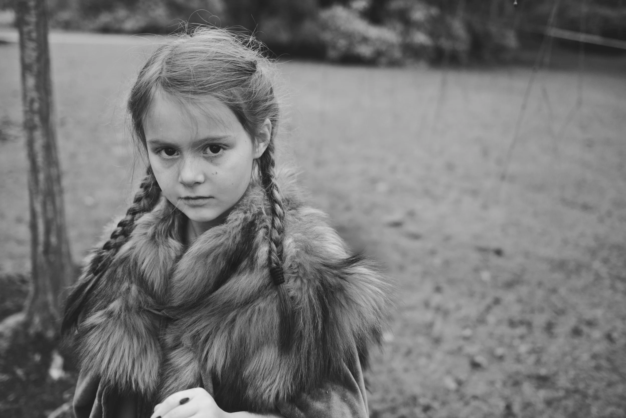 A young girl with braided hair and a fur coat looks directly at the camera in an outdoor setting with blurred trees and the ground in the background.