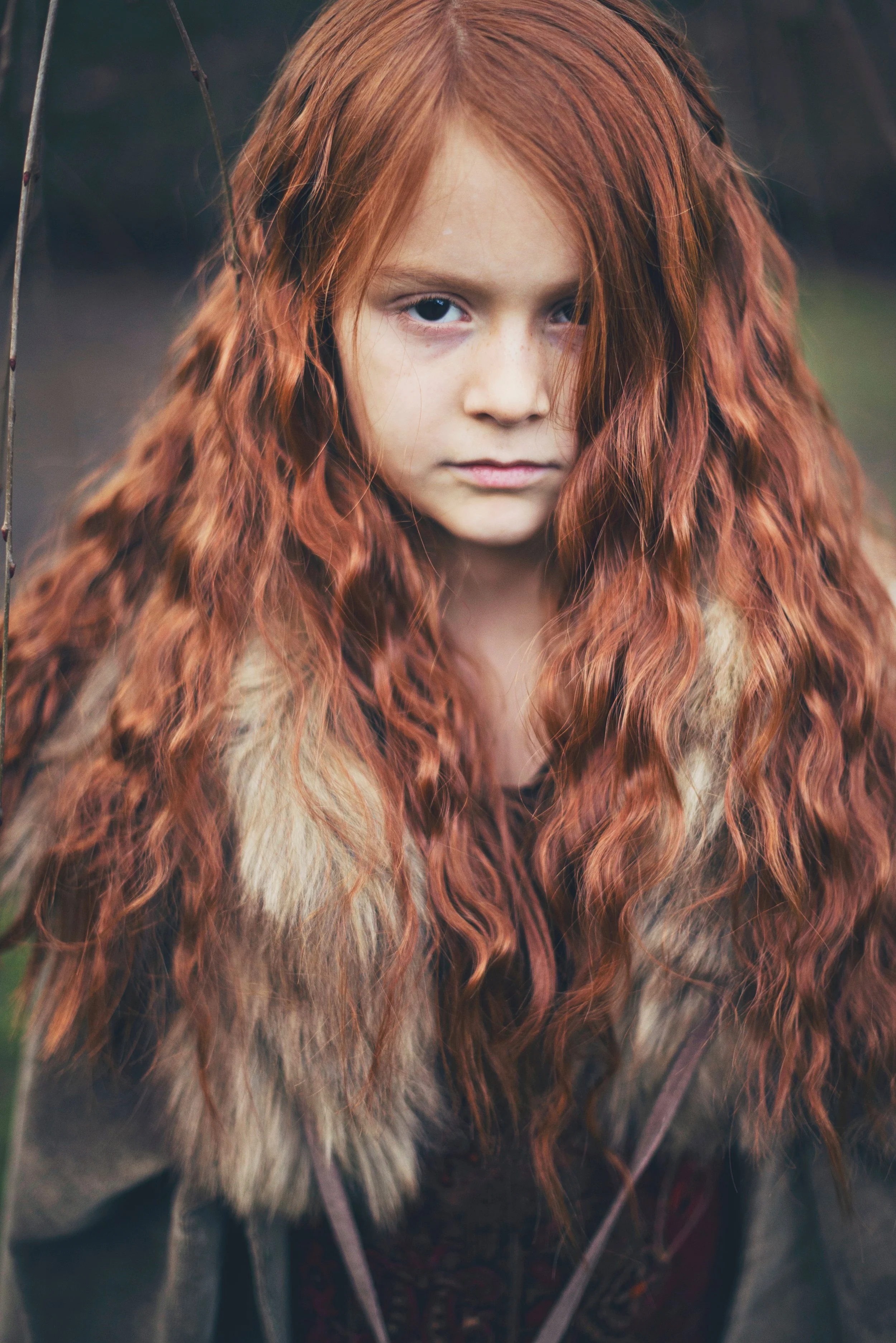 Close-up of a young girl with long, curly red hair, light skin, and blue eyes wearing a fur cloak, outdoors in a natural setting.