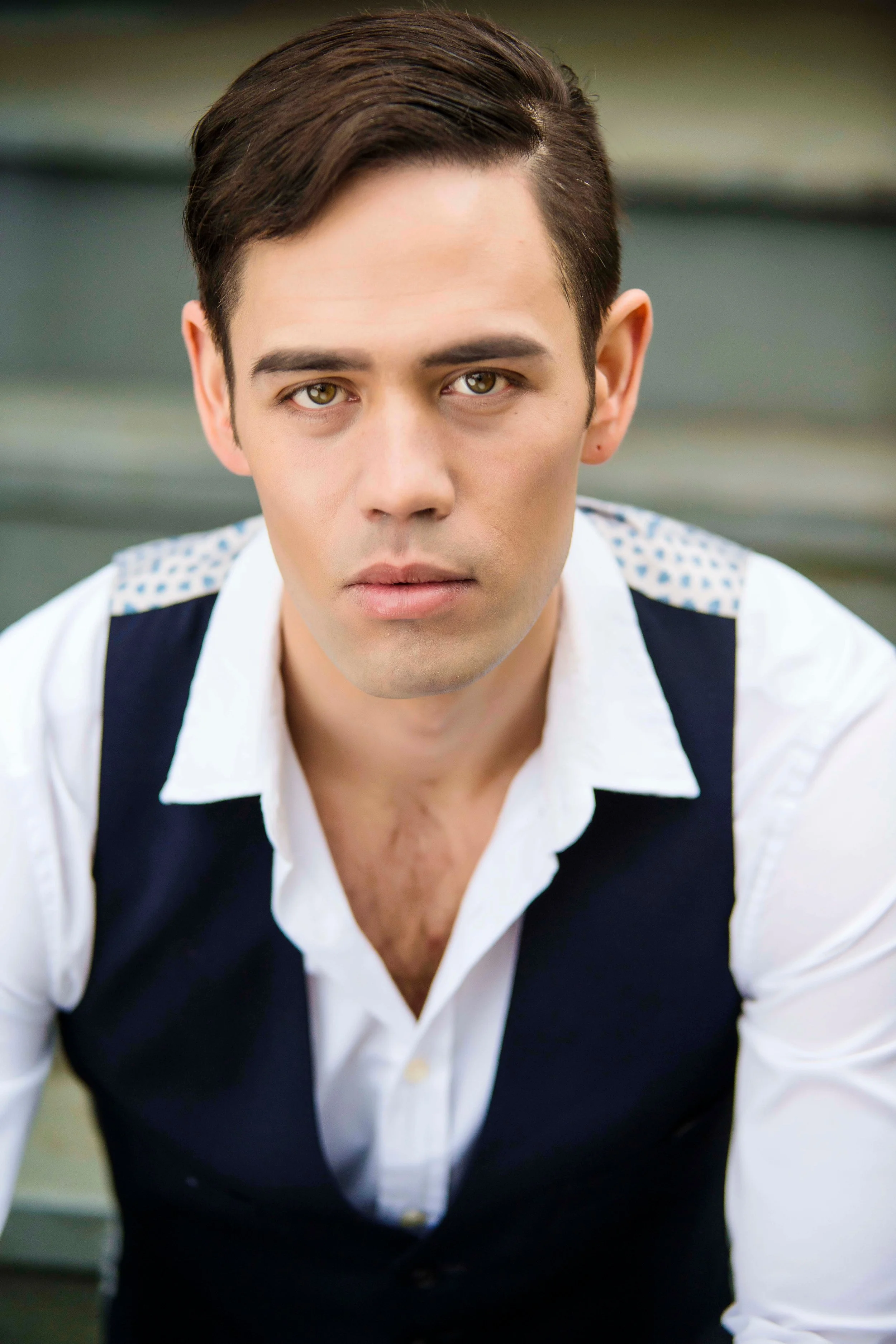 Close-up of a young man with brown hair and hazel eyes wearing a white shirt and black vest, looking directly at the camera with a neutral expression.