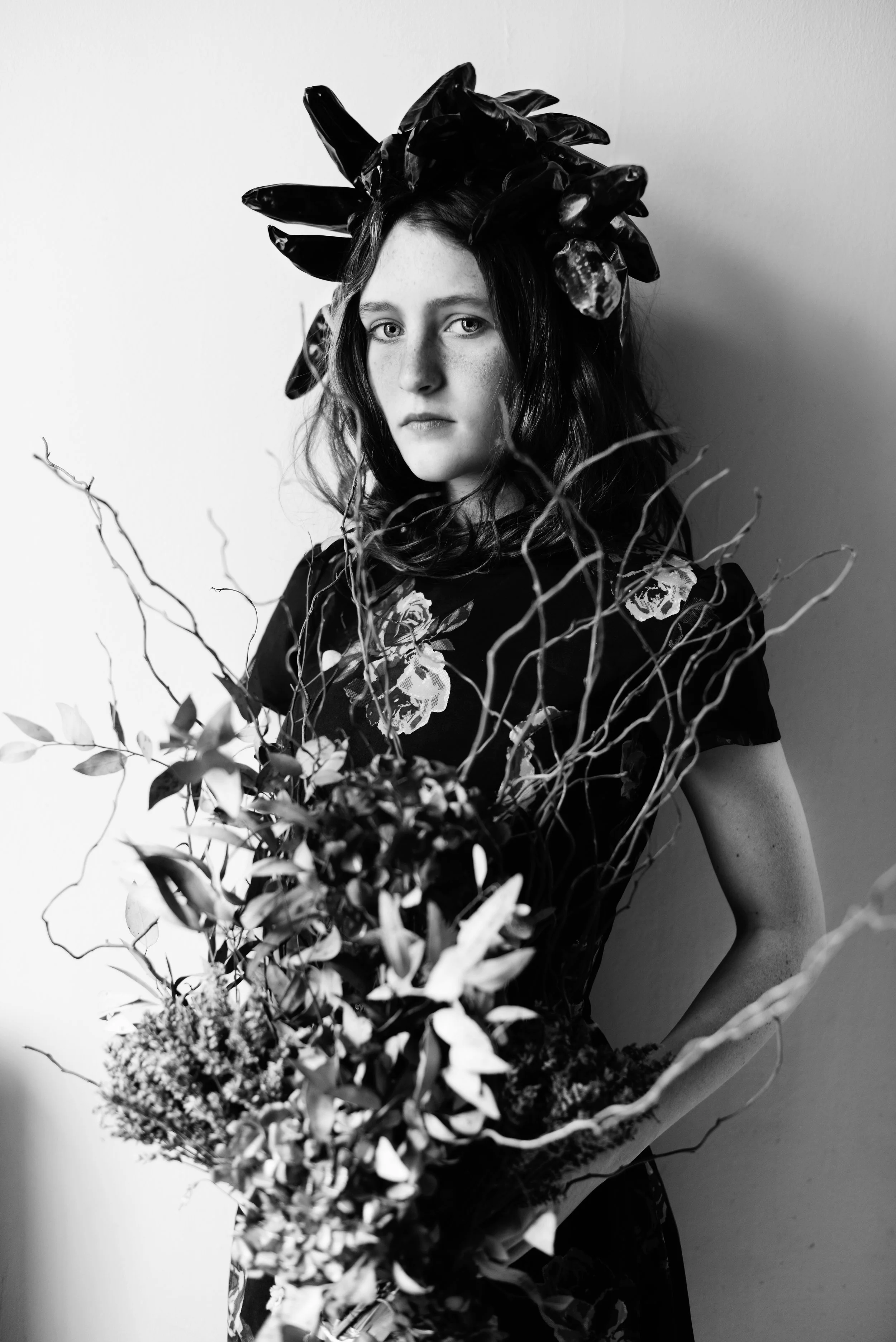 Black and white photograph of a young woman with curly hair, wearing a floral patterned dress, holding a bouquet of flowers and branches, standing against a plain wall, with a large leafy headpiece on her head.
