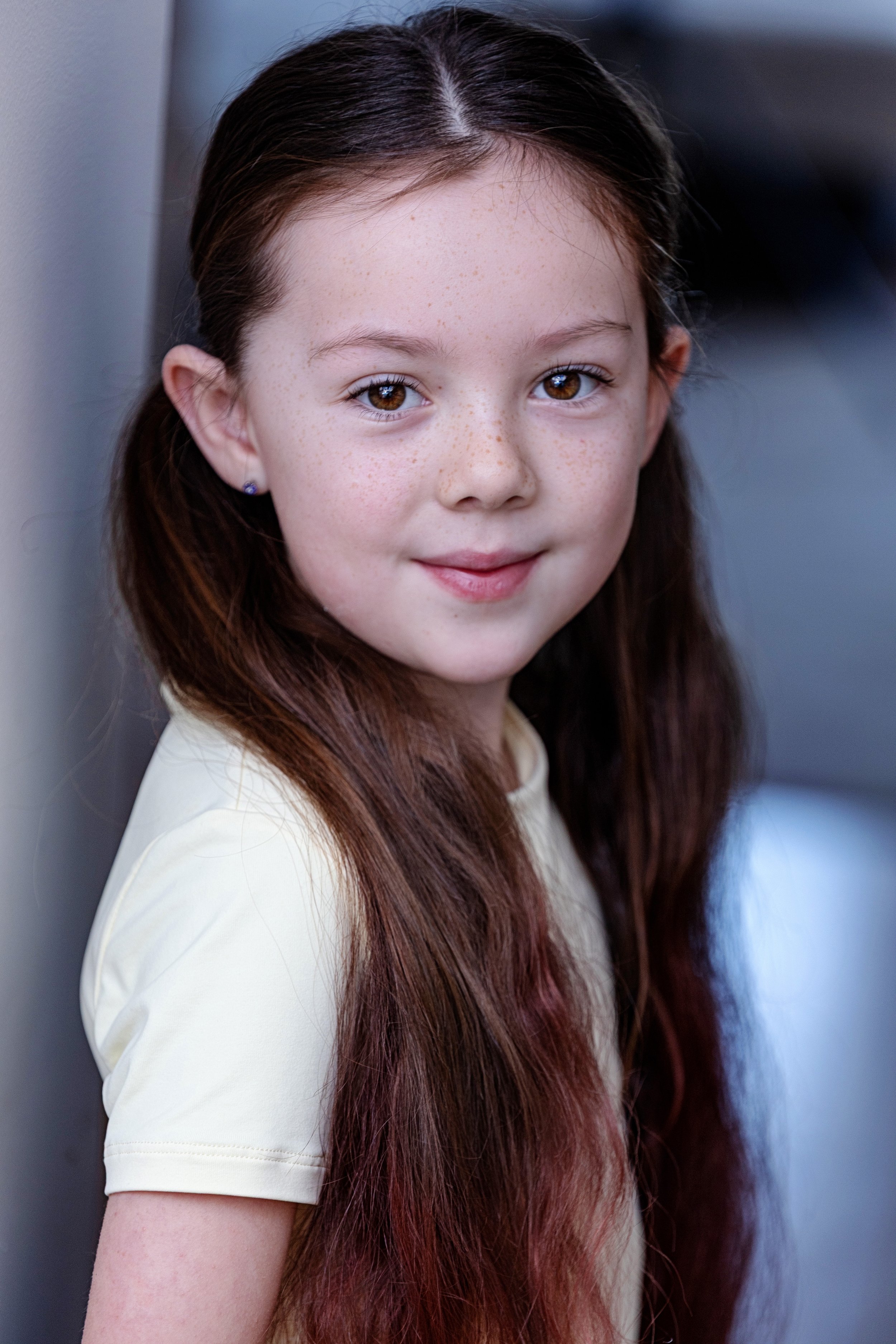 Close-up of a young girl with long, reddish-brown hair, wearing a light-colored shirt, standing against a gray wall.