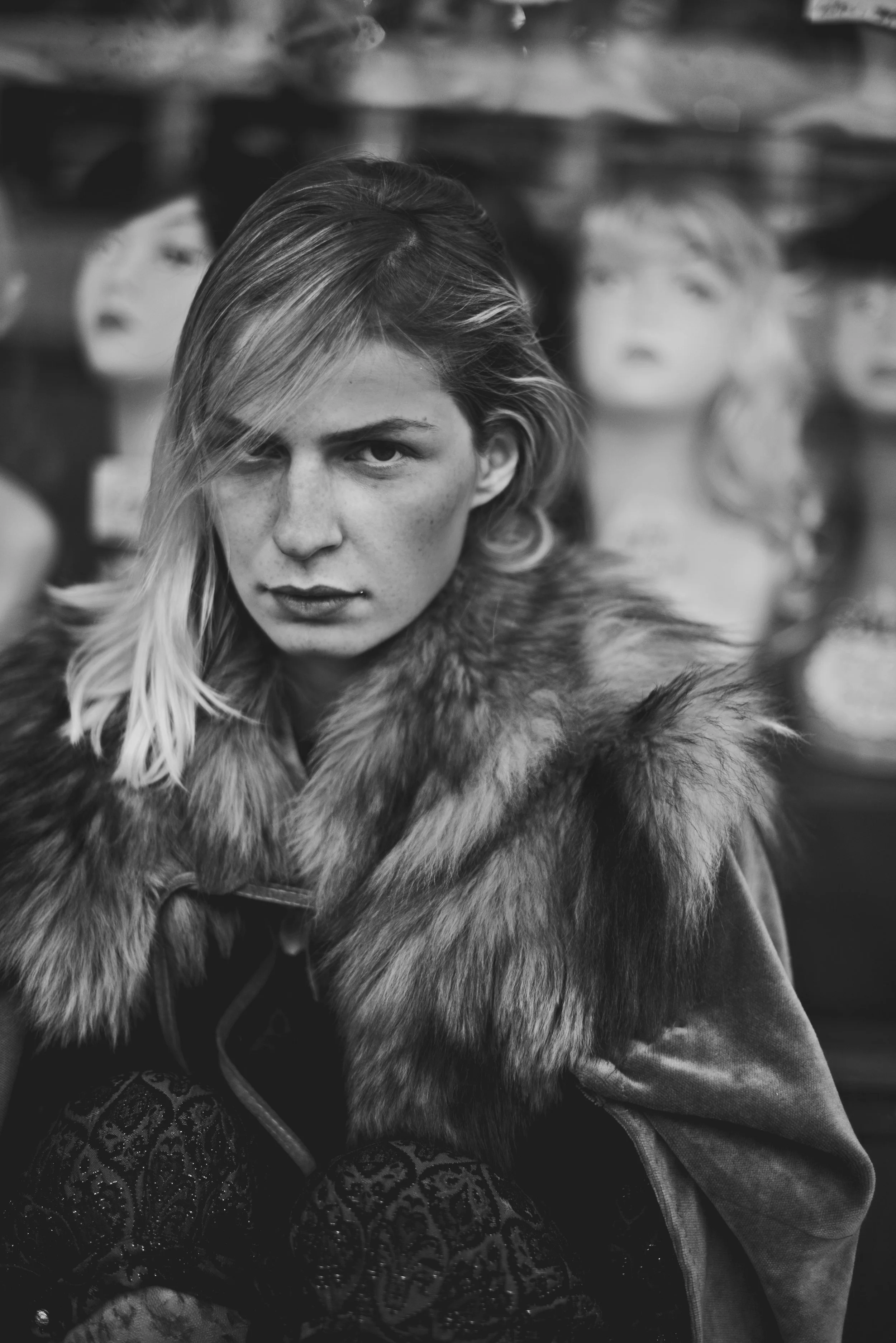 Black and white photograph of a woman with long hair, wearing a fur coat, sitting in front of mannequin heads with wigs.