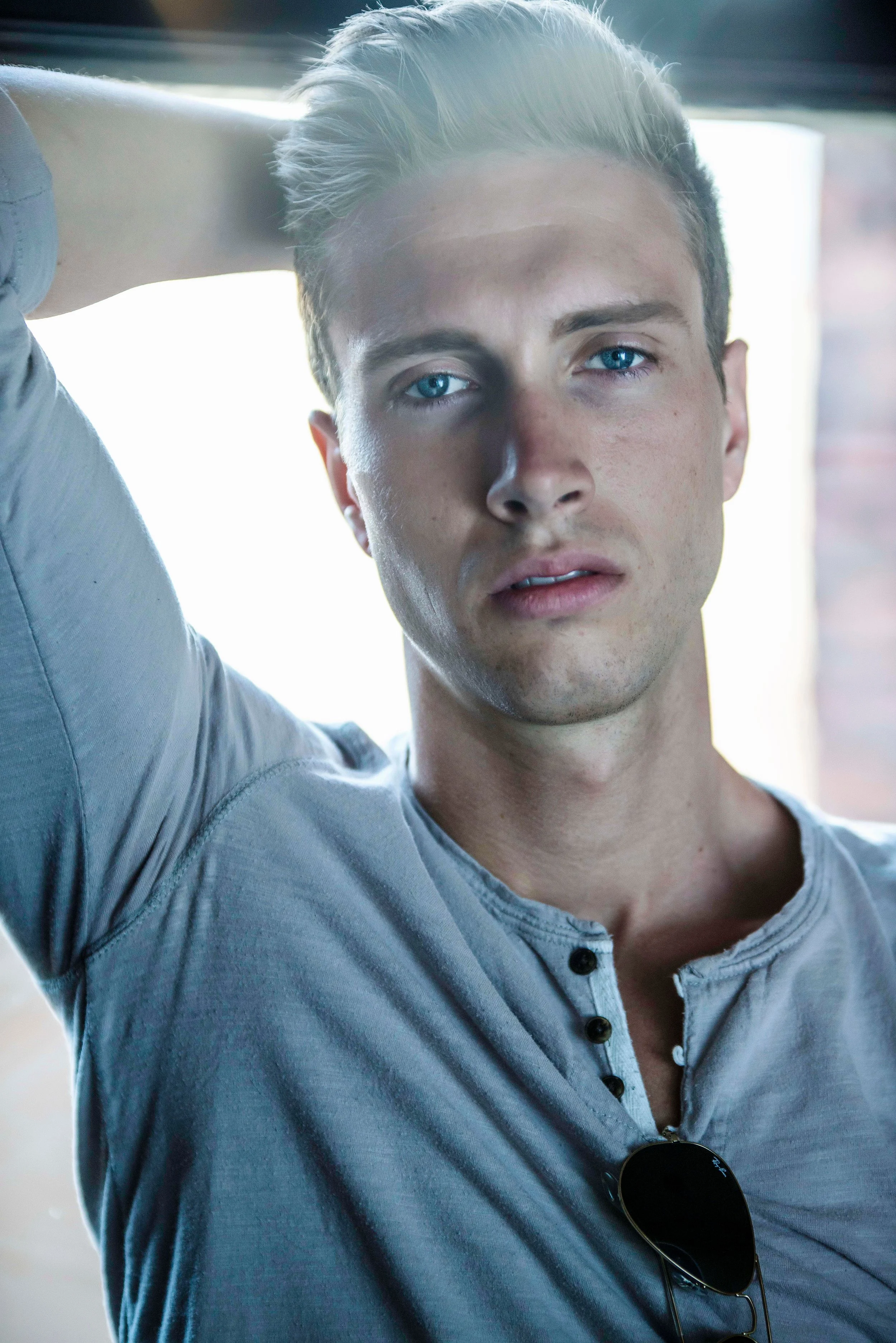 Close-up of a young man with light skin, blue eyes, and blonde hair, wearing a light blue shirt with dark buttons. He has sunglasses hanging from his shirt and is posing indoors with his arm resting behind his head, looking into the camera.