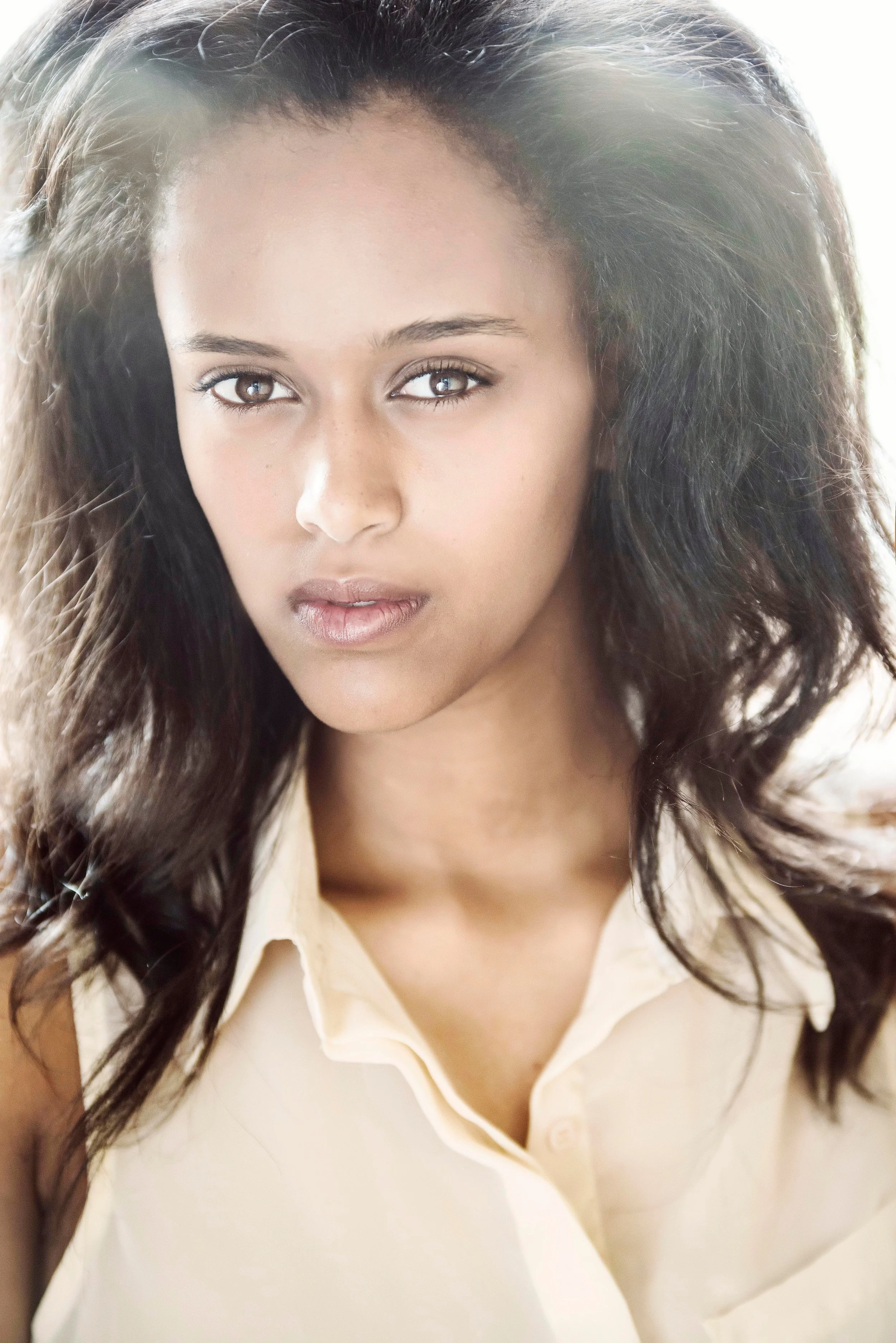 A close-up portrait of a young woman with long, wavy dark hair and brown eyes, wearing a light-colored collared shirt.