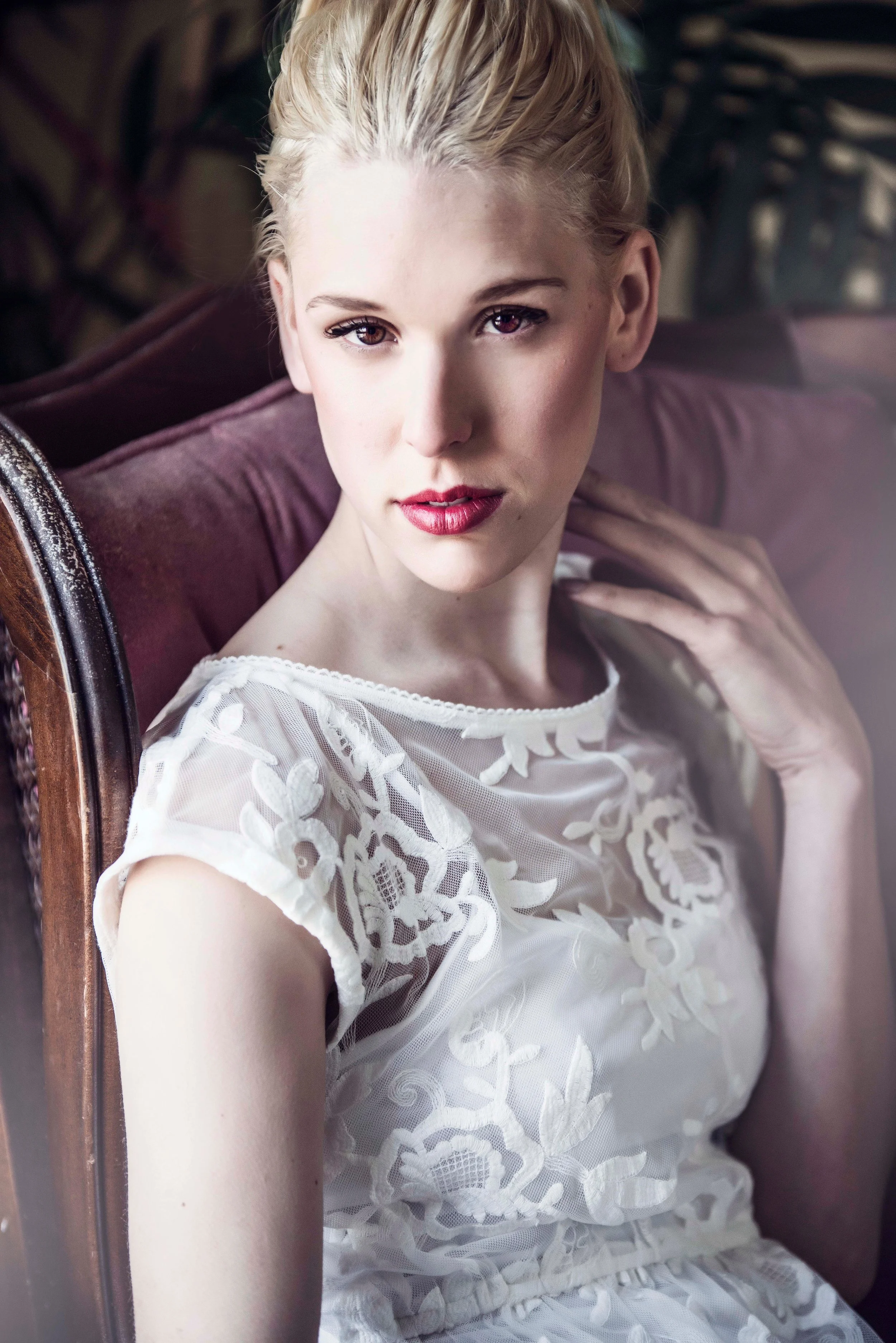 A young woman with blonde hair and fair skin is sitting on a vintage wooden chair with a pink cushion. She is wearing a white, floral lace dress and looking directly at the camera with a slight smile.