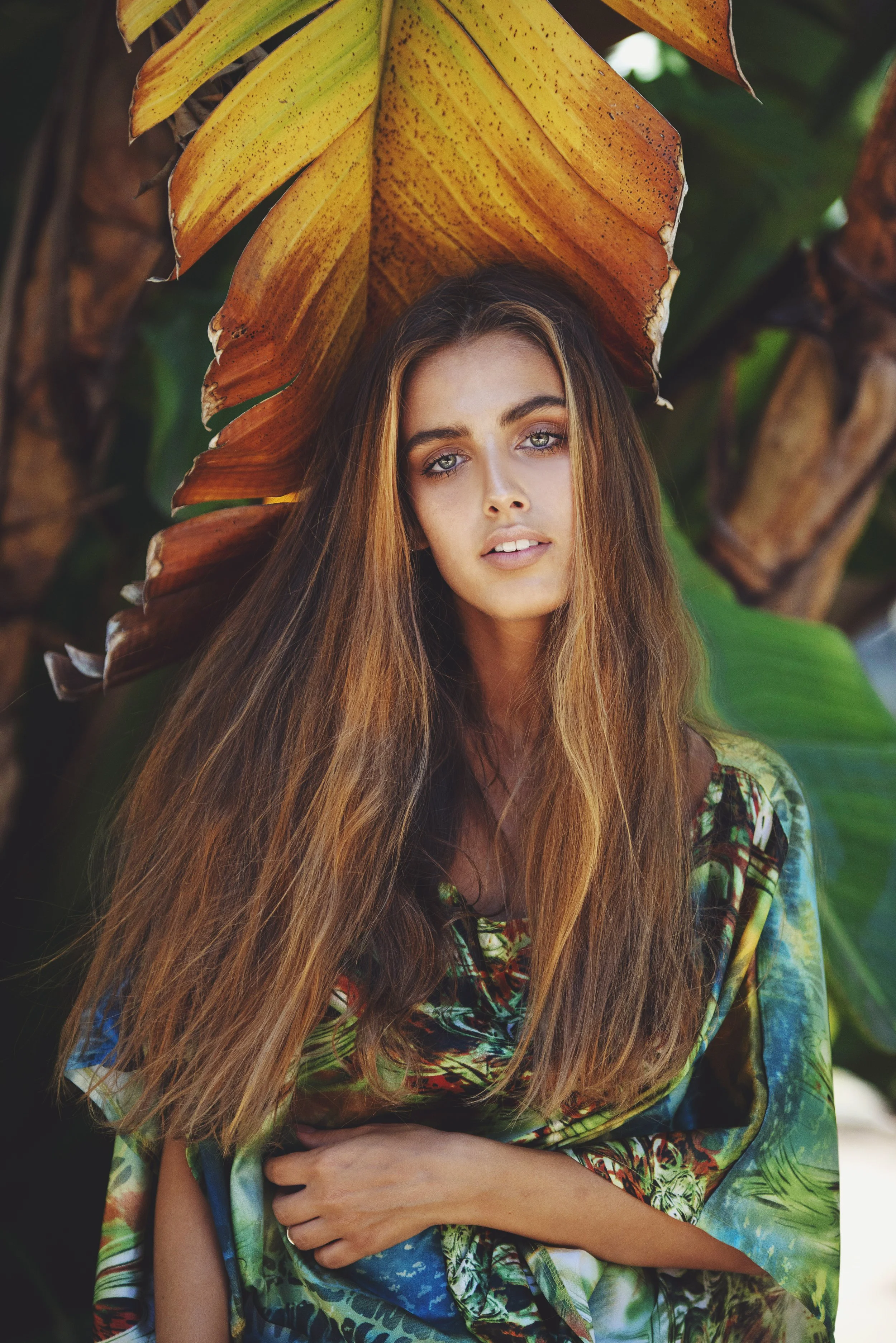 A young woman with long, reddish-brown hair and blue eyes, wearing a colorful, tropical-print top, standing outdoors with large green and brown tropical leaves in the background.