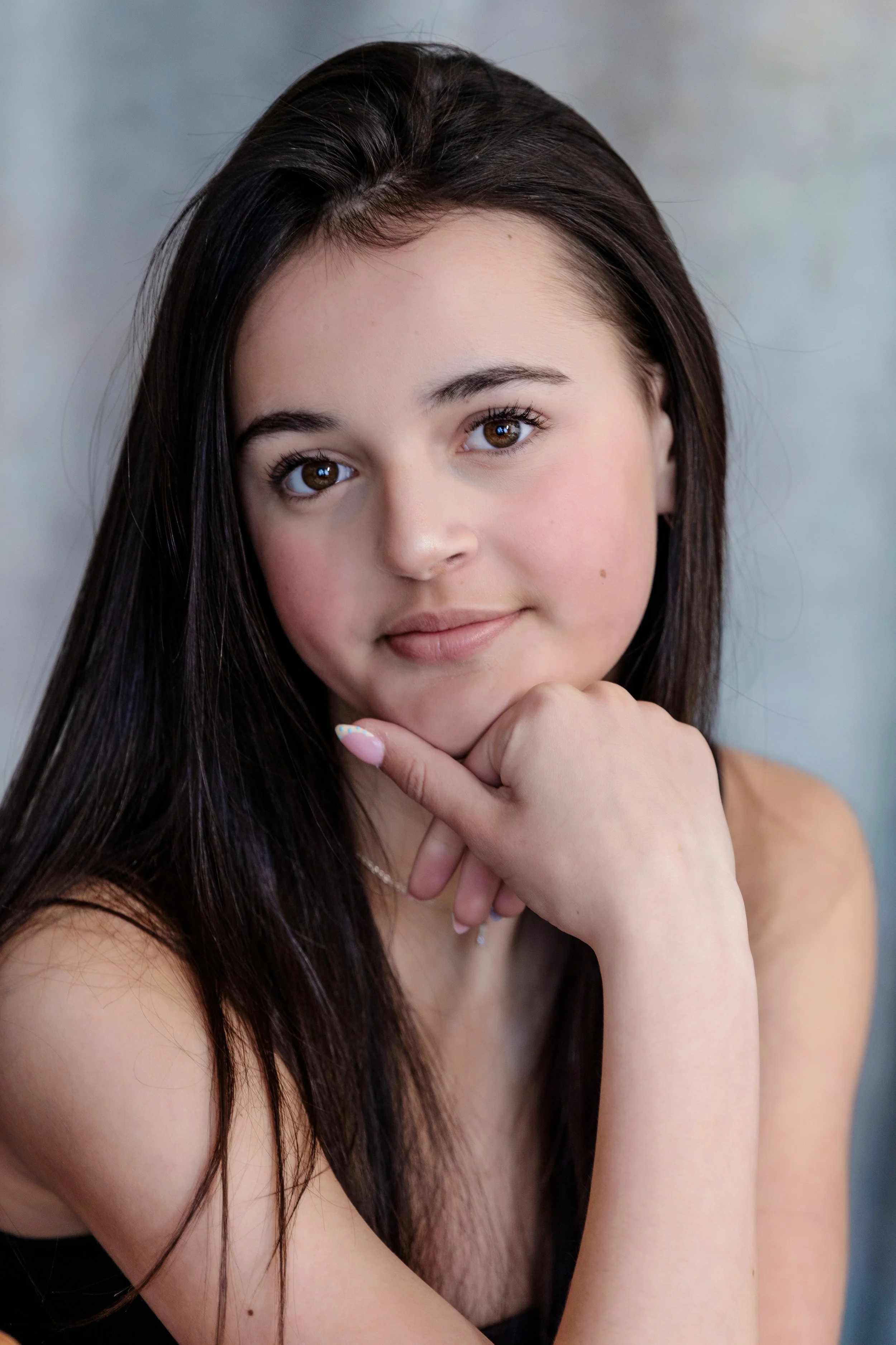 Close-up portrait of a young woman with long dark hair, brown eyes, and a gentle smile, resting her chin on her hand.