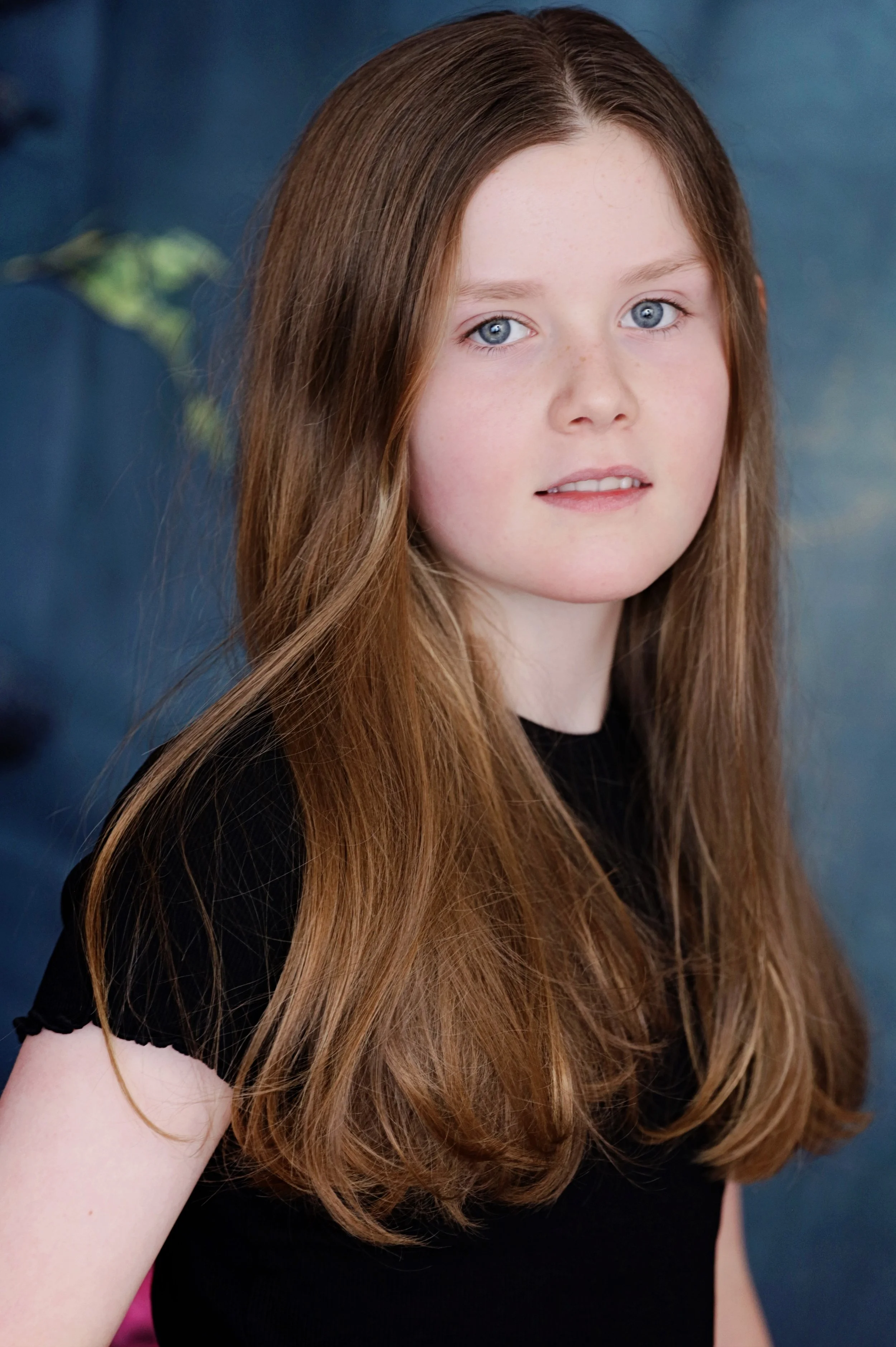 Close-up of a girl with long, wavy red hair and blue eyes wearing a black shirt, against a blue background.