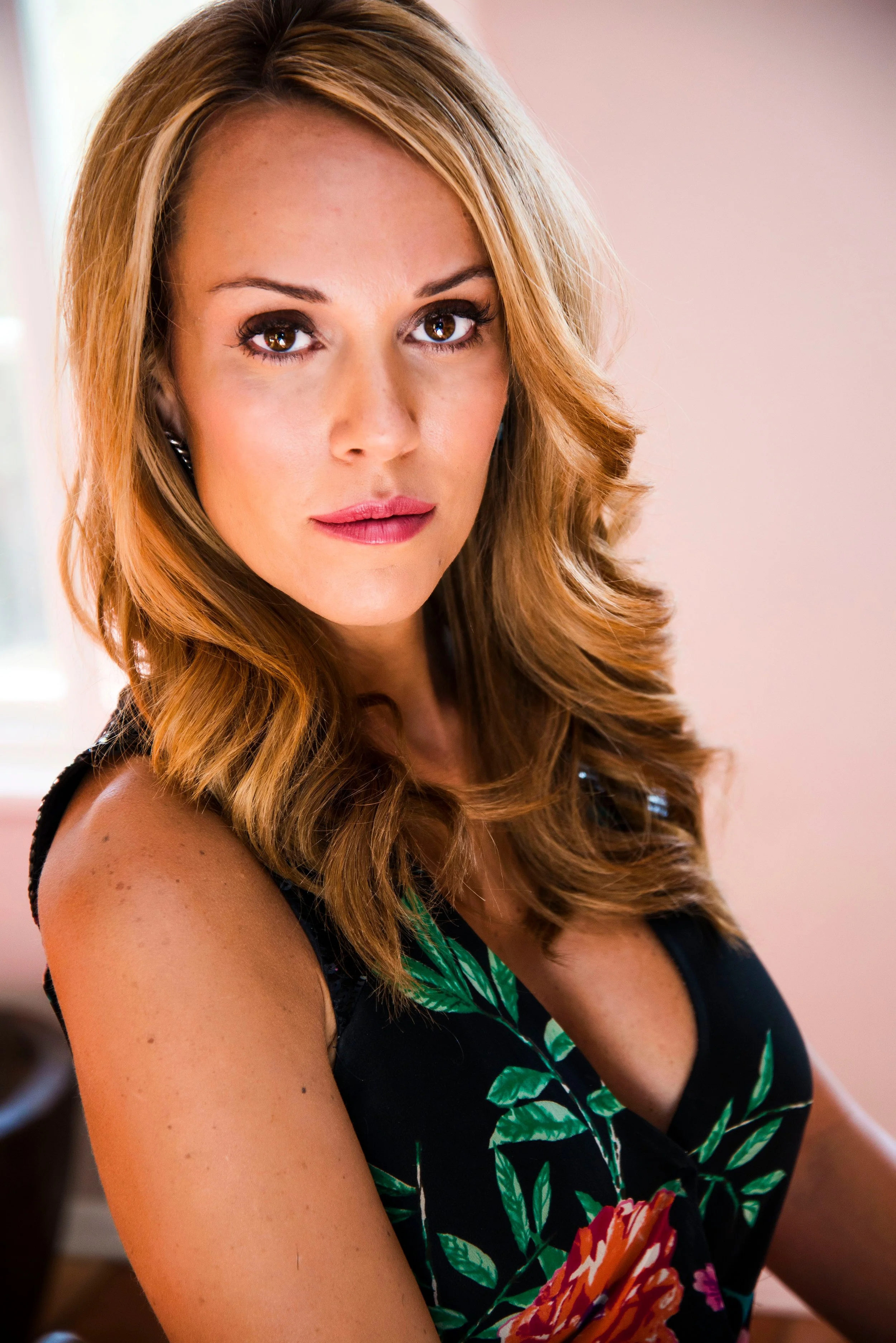 Close-up of a woman with wavy, reddish-brown hair, brown eyes, and light makeup, wearing a sleeveless black dress with a colorful floral pattern, sitting indoors with a soft pink background.