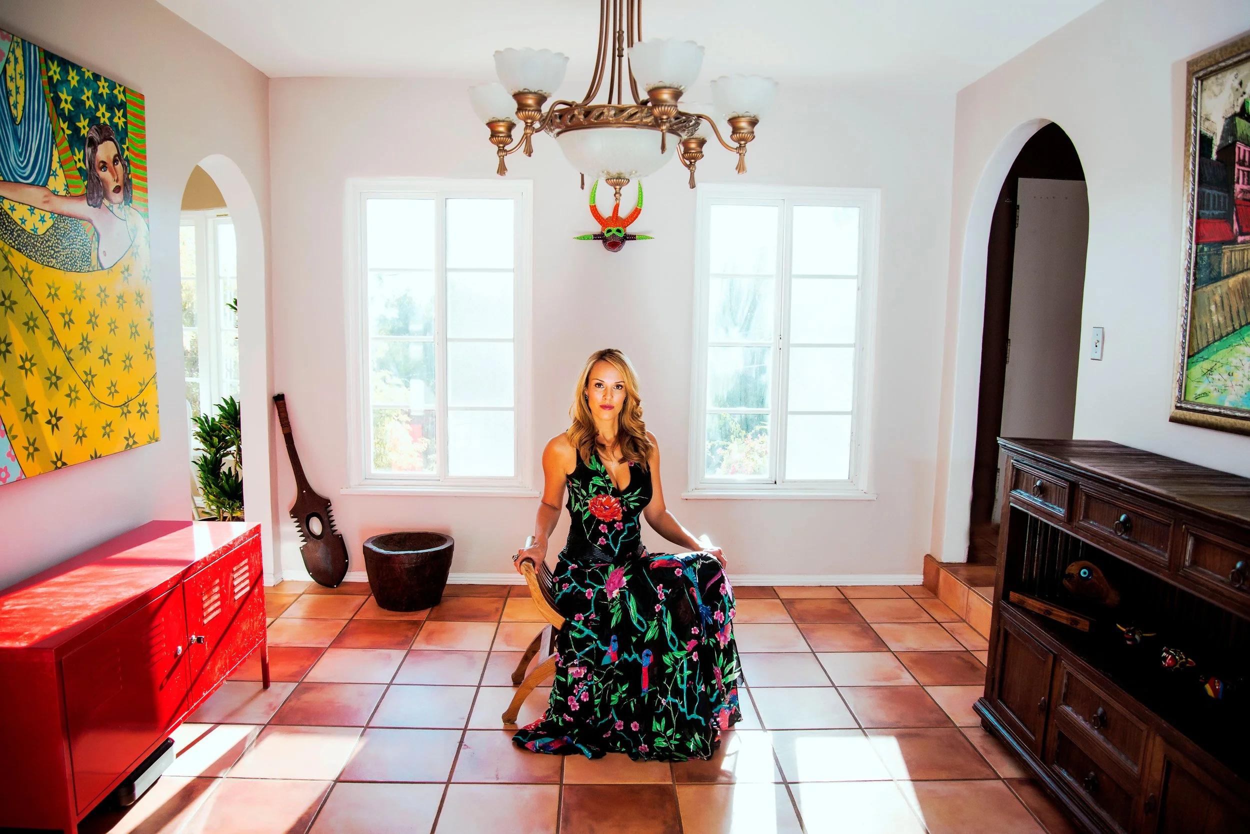 A woman with wavy blonde hair sitting on a wooden chair in a bright room, wearing a black dress with pink, green, and blue floral patterns. The room has white walls, large windows, a vintage chandelier, colorful artwork, a red cabinet, and other furn