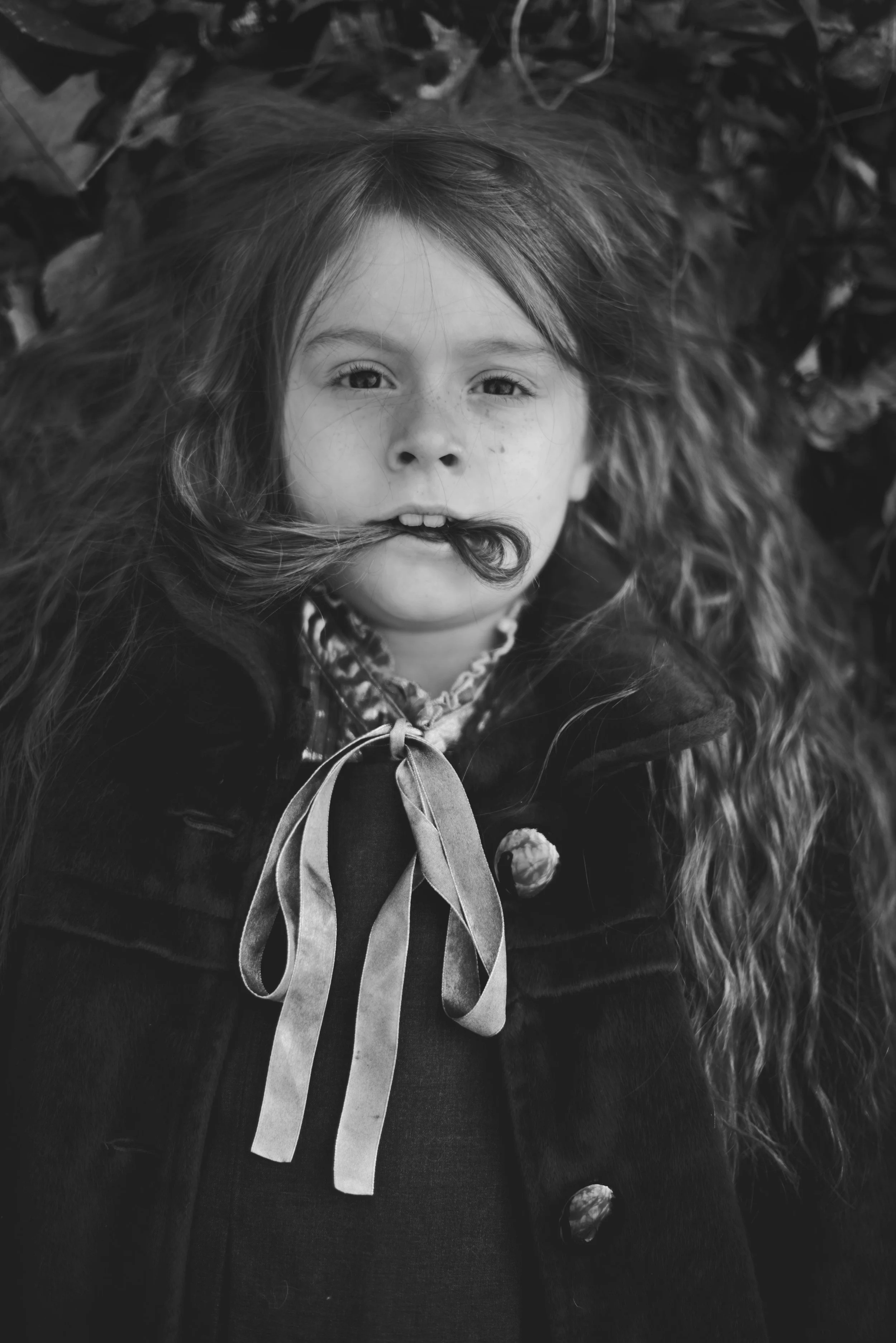 Black and white photo of a young girl lying on the ground with leaves around her. She has long hair, some strands in a curl, and her eyes are open, looking up. She is wearing a coat with a fur-lined hood and a patterned shirt with a ribbon tie.