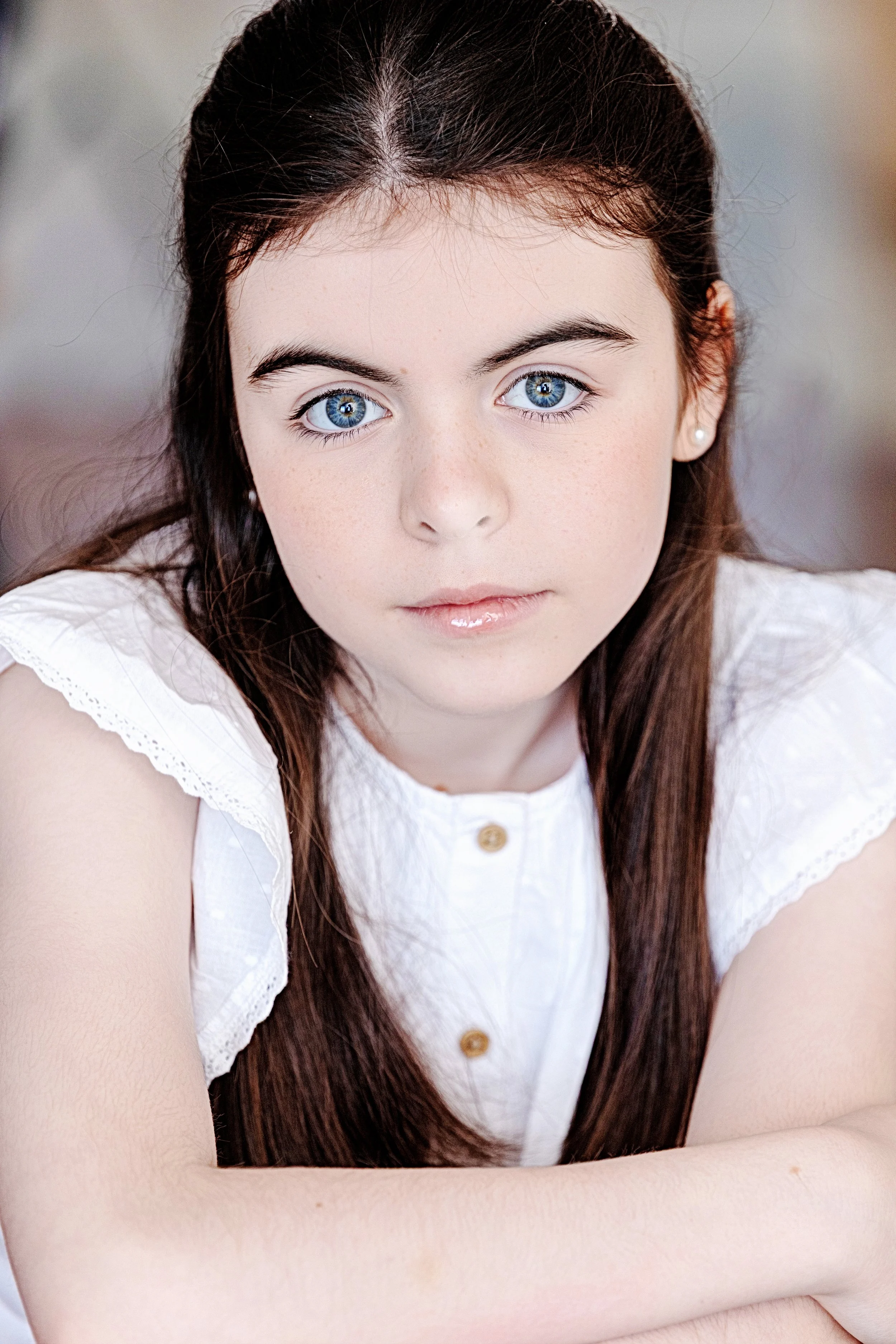 A young girl with long brown hair and blue eyes wearing a white sleeveless top with lace trim and pearl earrings, looking directly at the camera.