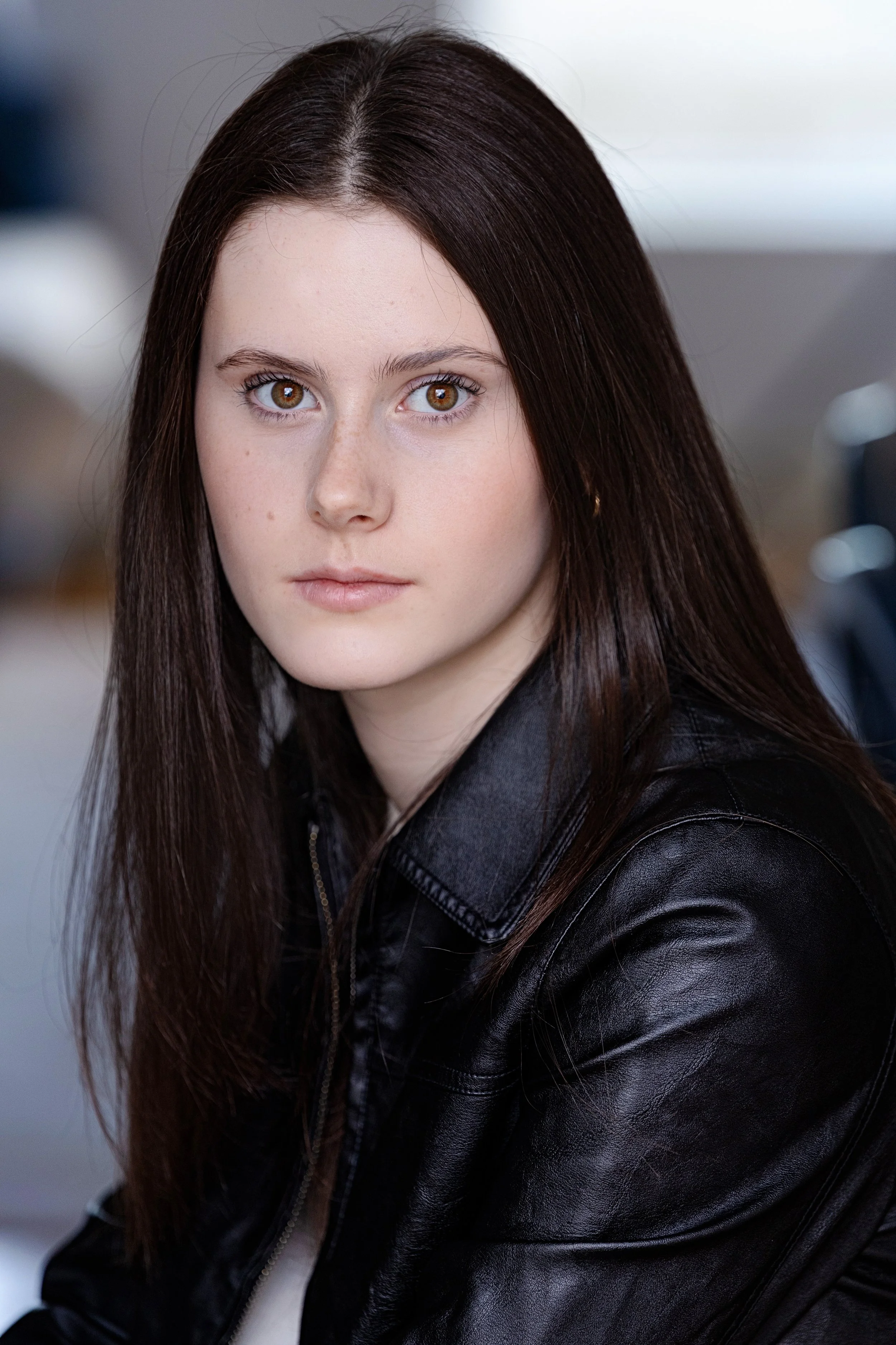 Close-up of a young woman with long brown hair, hazel eyes, and light skin, wearing a black leather jacket, looking directly at the camera with a neutral expression.