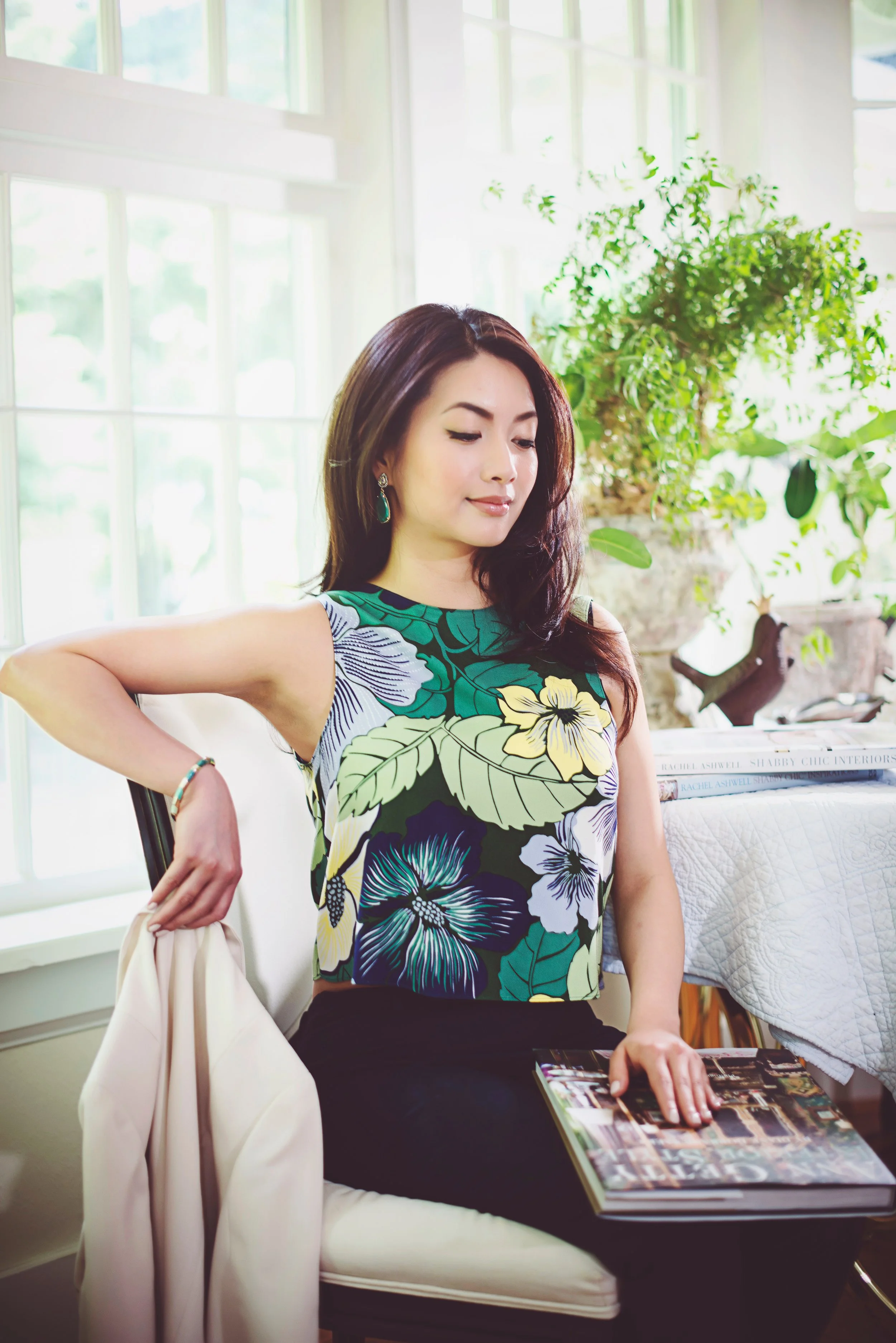 A woman seated indoors near a large window with sunlight, wearing a floral sleeveless top, earrings, and a bracelet, with a large potted plant behind her. She is holding a magazine on her lap and has a white handbag hanging on the chair.