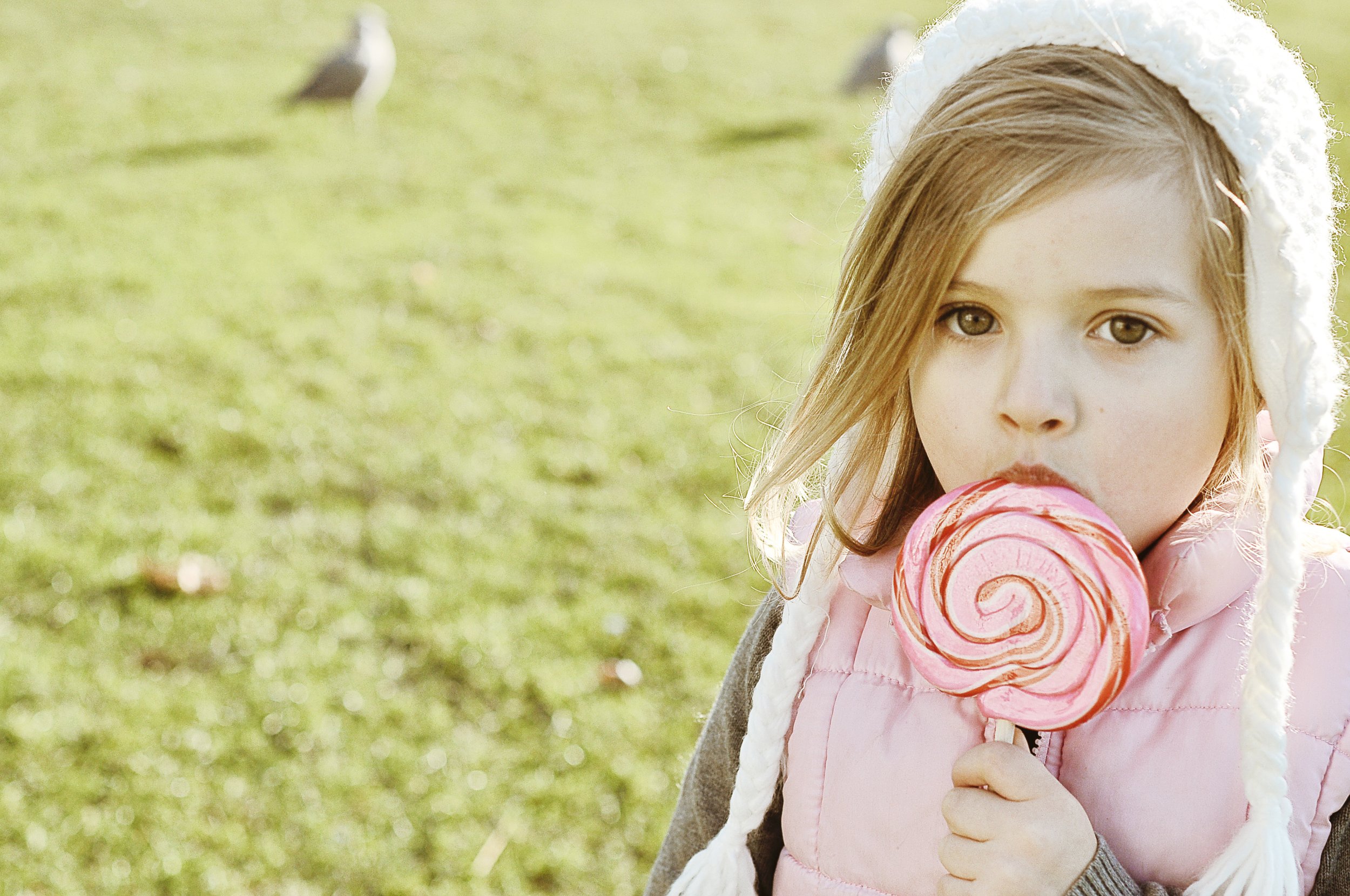 A young girl with light brown hair and hazel eyes holding a pink and white swirled lollipop outdoors on a sunny day, with a grassy field and birds in the background.