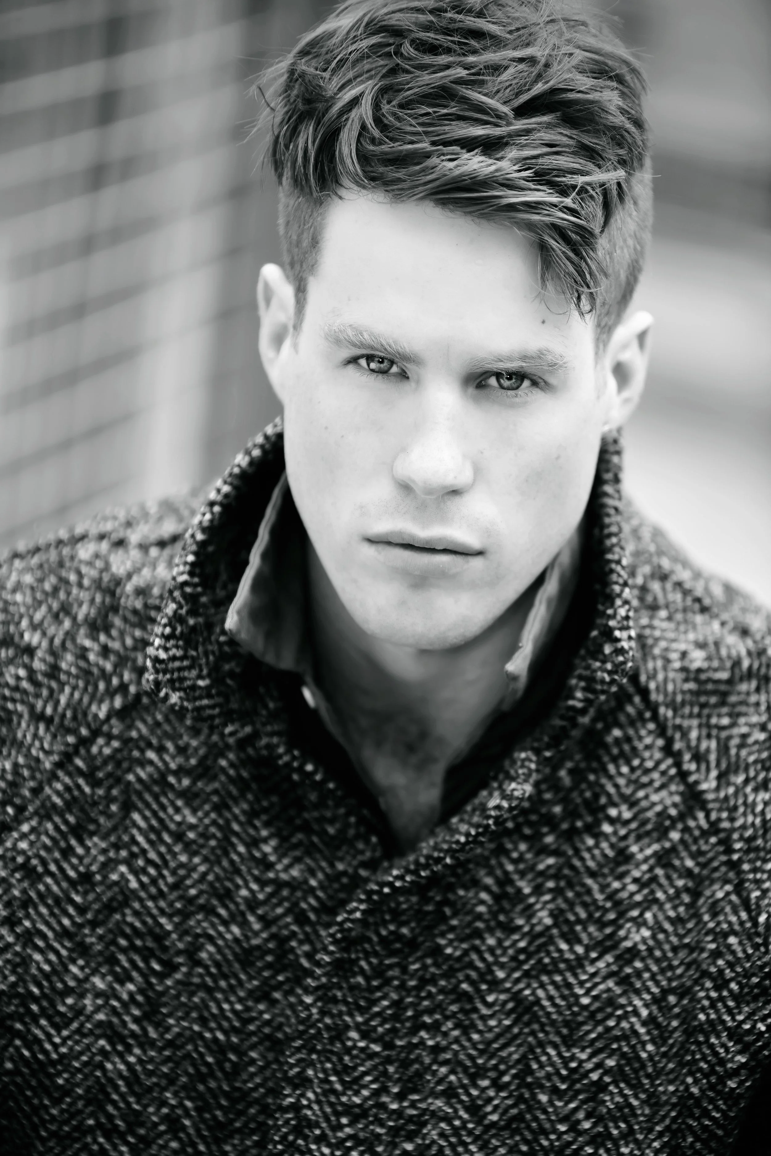 Black and white close-up portrait of a young man with styled hair and intense gaze, wearing a textured coat and collared shirt.