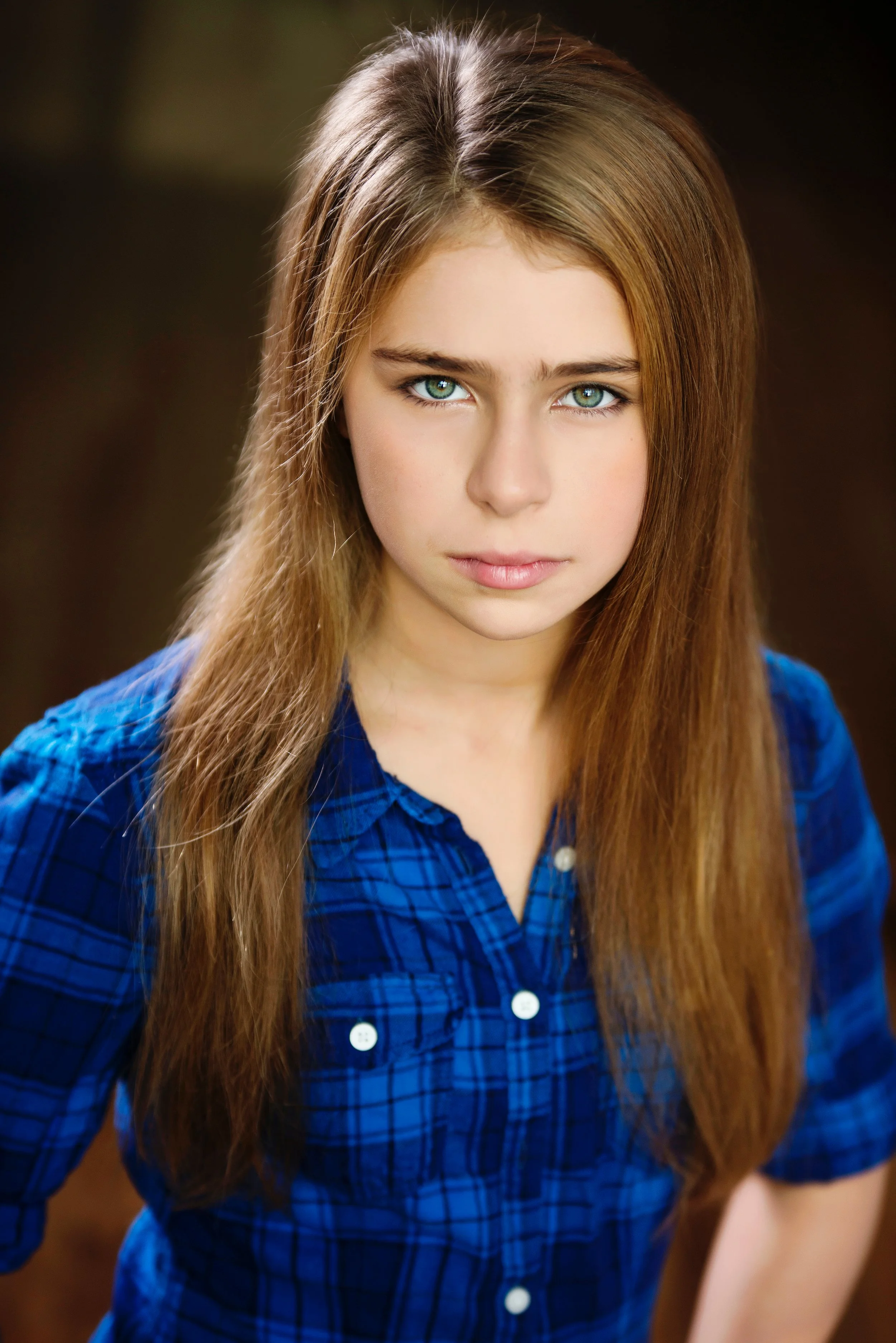 Close-up portrait of a young woman with long, straight, light brown hair and blue eyes. She is wearing a blue plaid shirt and looking directly at the camera.