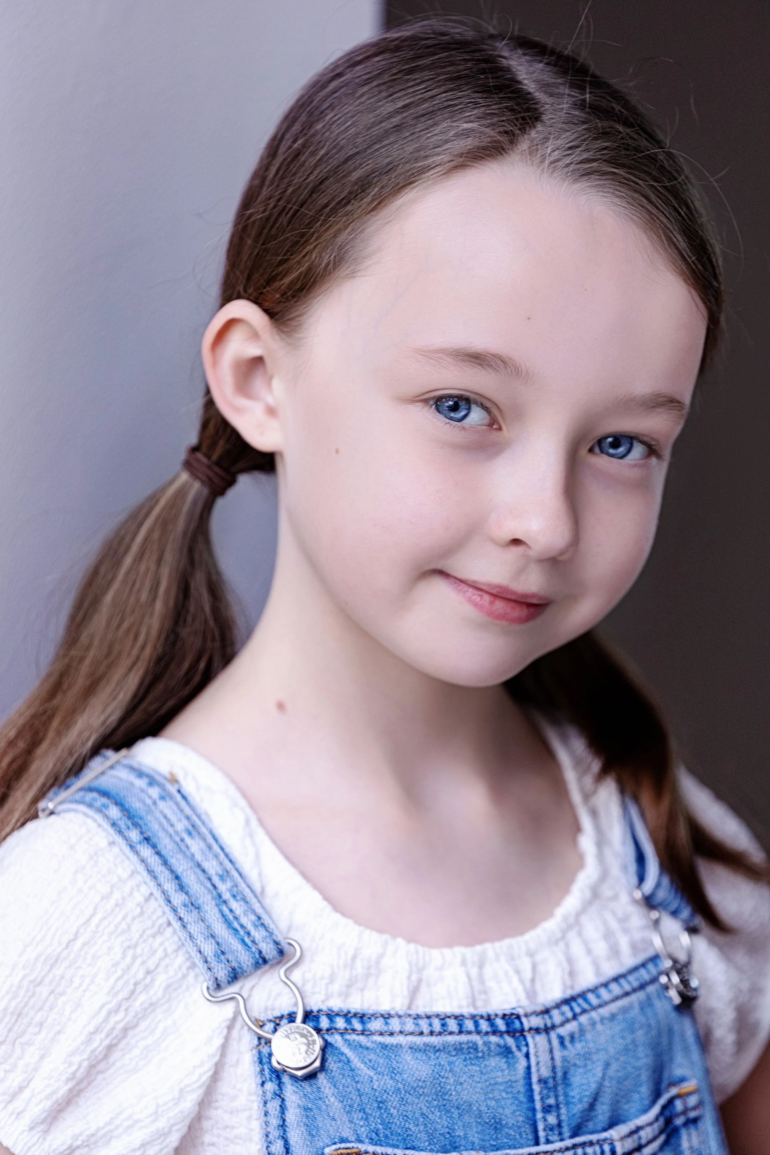 Close-up of a young girl with blue eyes and brown hair in pigtails, wearing a white shirt and denim overalls.