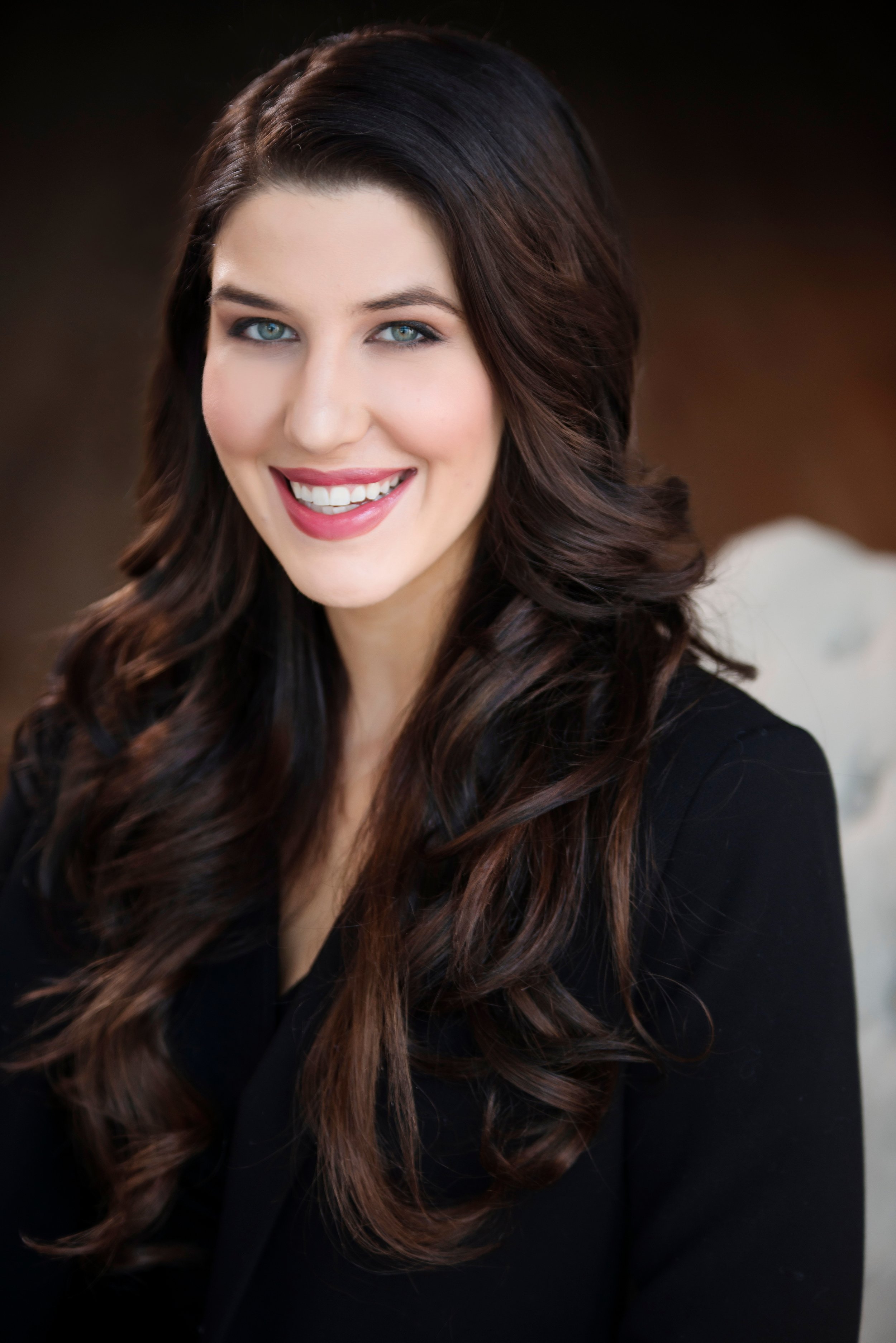 Close-up portrait of a smiling woman with long, wavy dark brown hair and blue eyes, wearing a black top, against a blurred dark background.