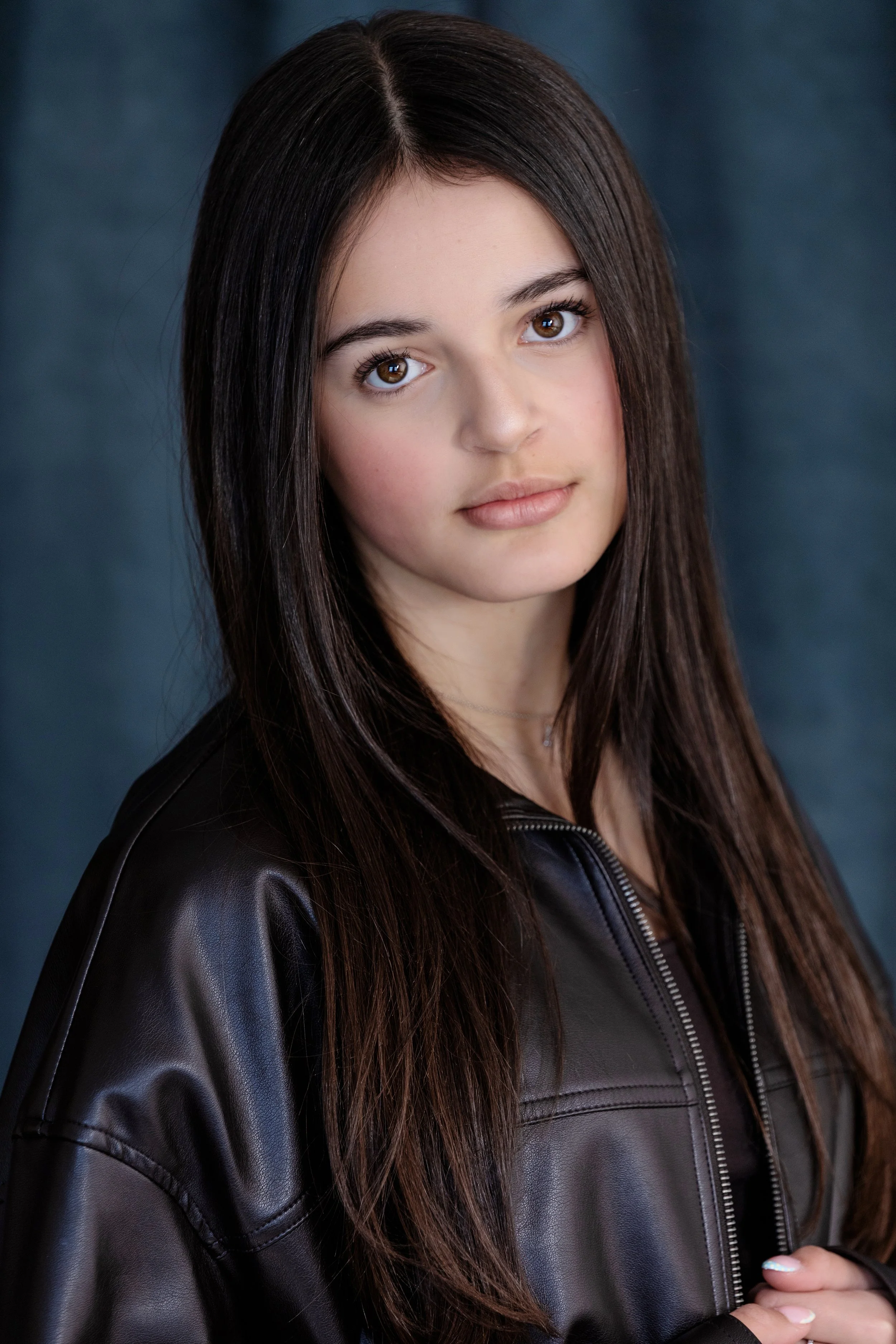 Portrait of a young woman with long dark hair wearing a black leather jacket, looking directly at the camera with a neutral expression, against a dark background.