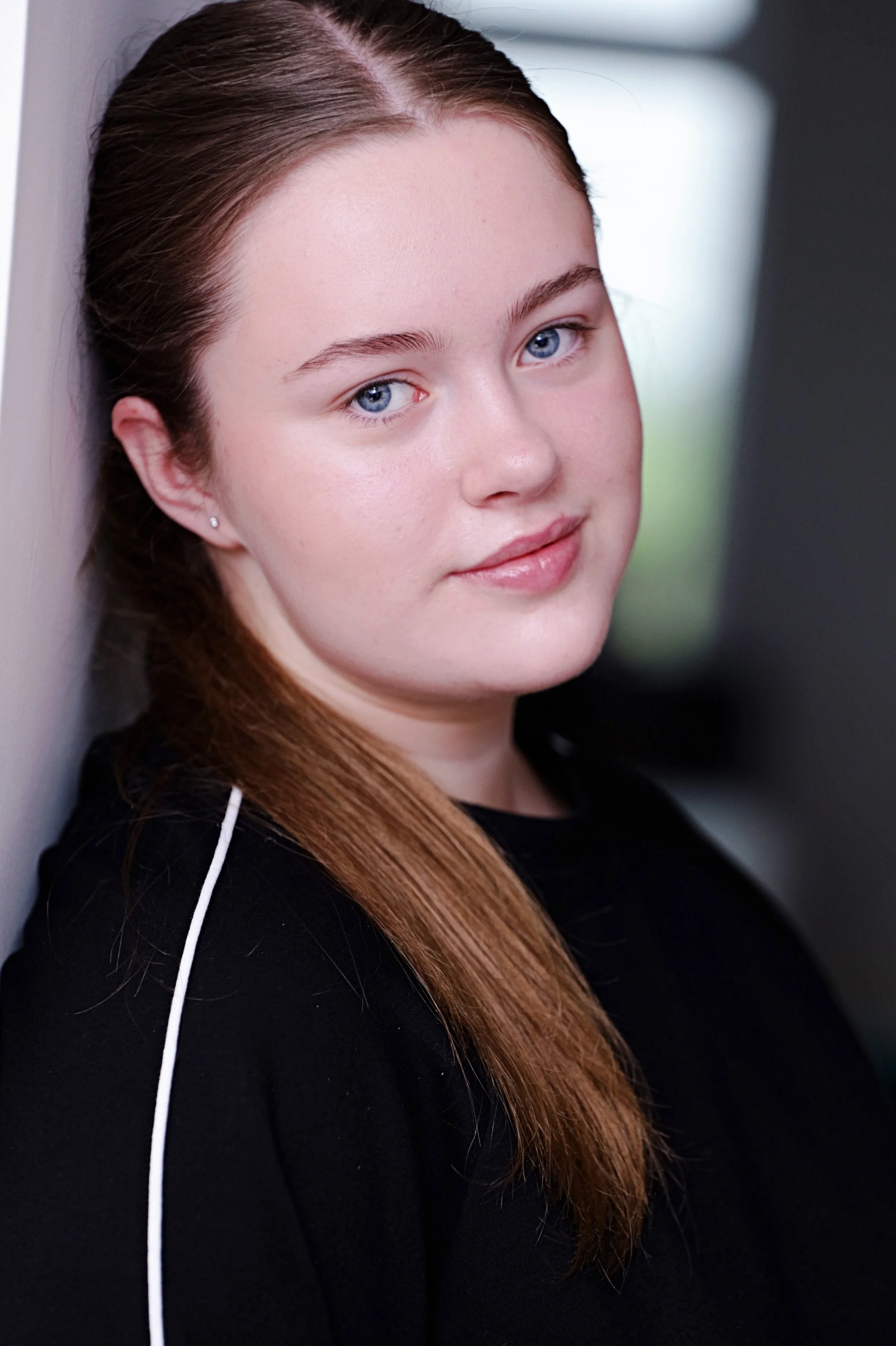 Close-up of a young woman with long brown hair, positioned sideways against a wall, looking directly at the camera with a slight smile, wearing a black top and a pearl earring.