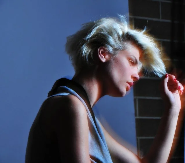 A woman with blonde, tousled hair sitting with her eyes closed, resting her head against a brick wall.