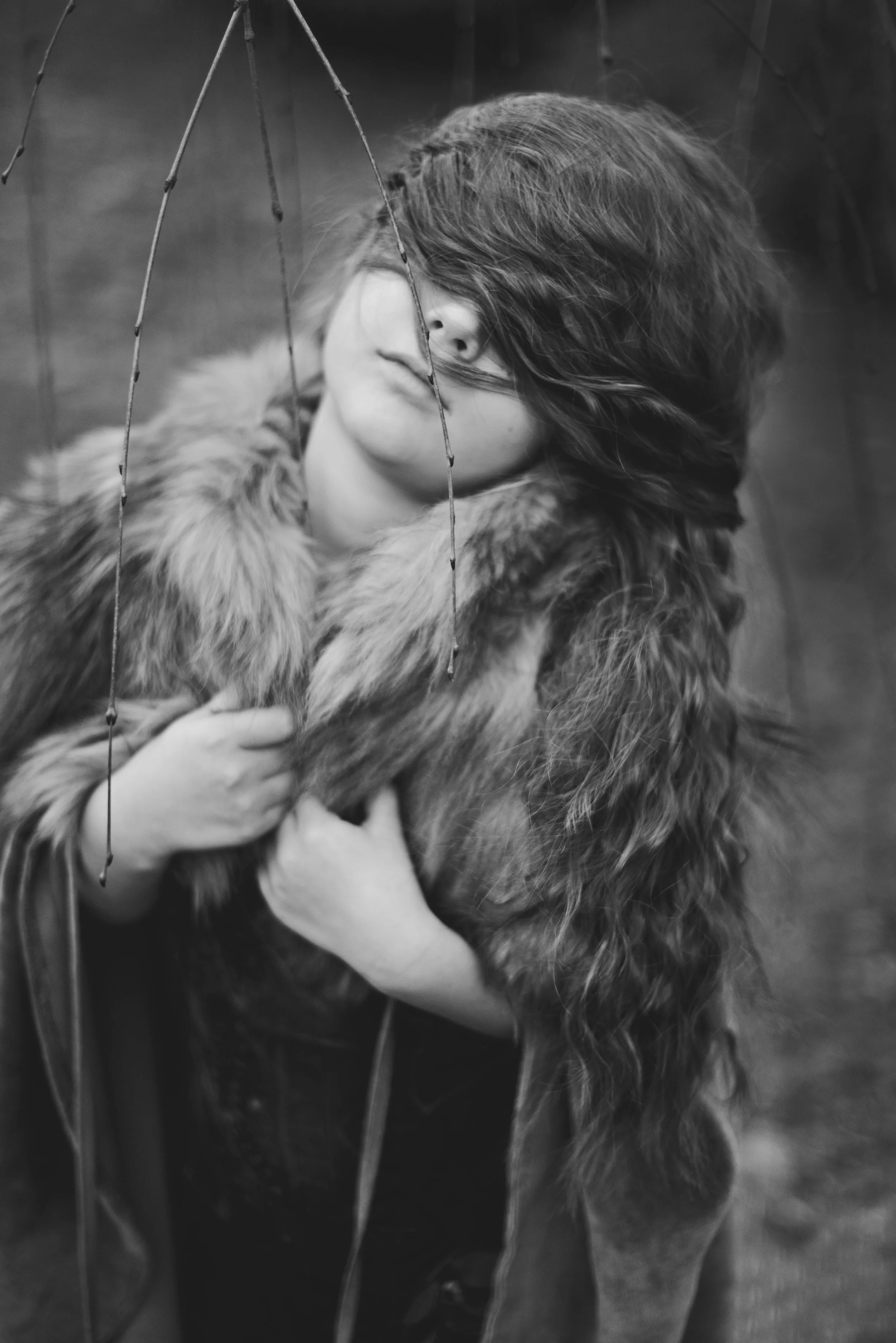 A person with long hair covering part of their face, wearing a fur-lined coat, holding a cat in their arms, with a blurred background.