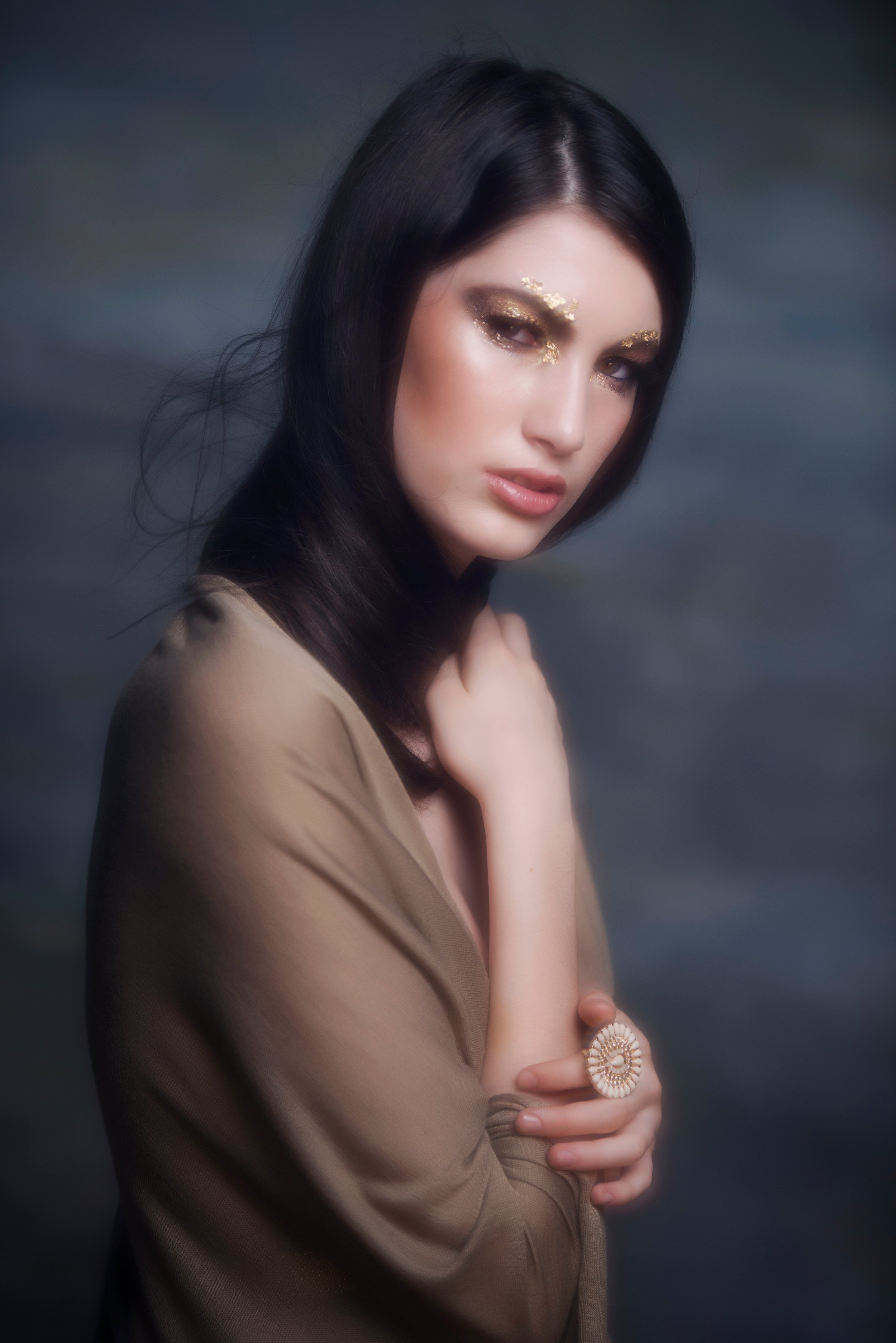 A portrait of a woman with dark hair, gold eye makeup, and a neutral expression, wearing a beige top and jewelry, with a dark cloudy background.