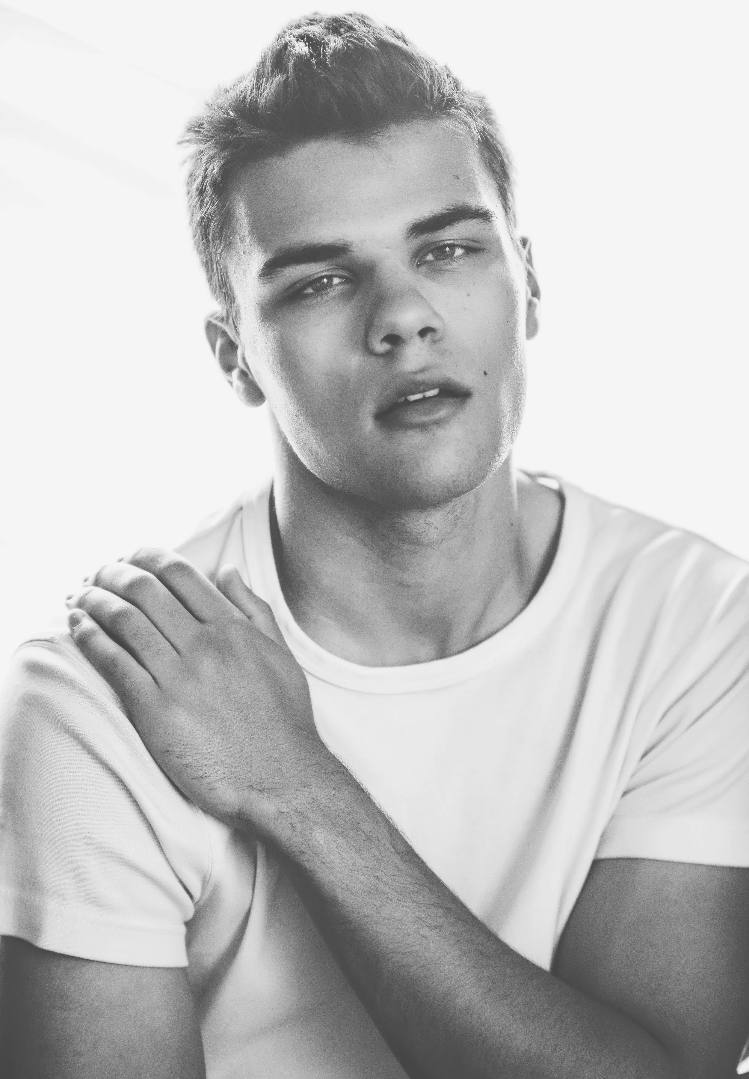 Black and white portrait of a young man with short hair, wearing a white T-shirt, looking into the camera with a serious expression, with one hand resting on his shoulder.