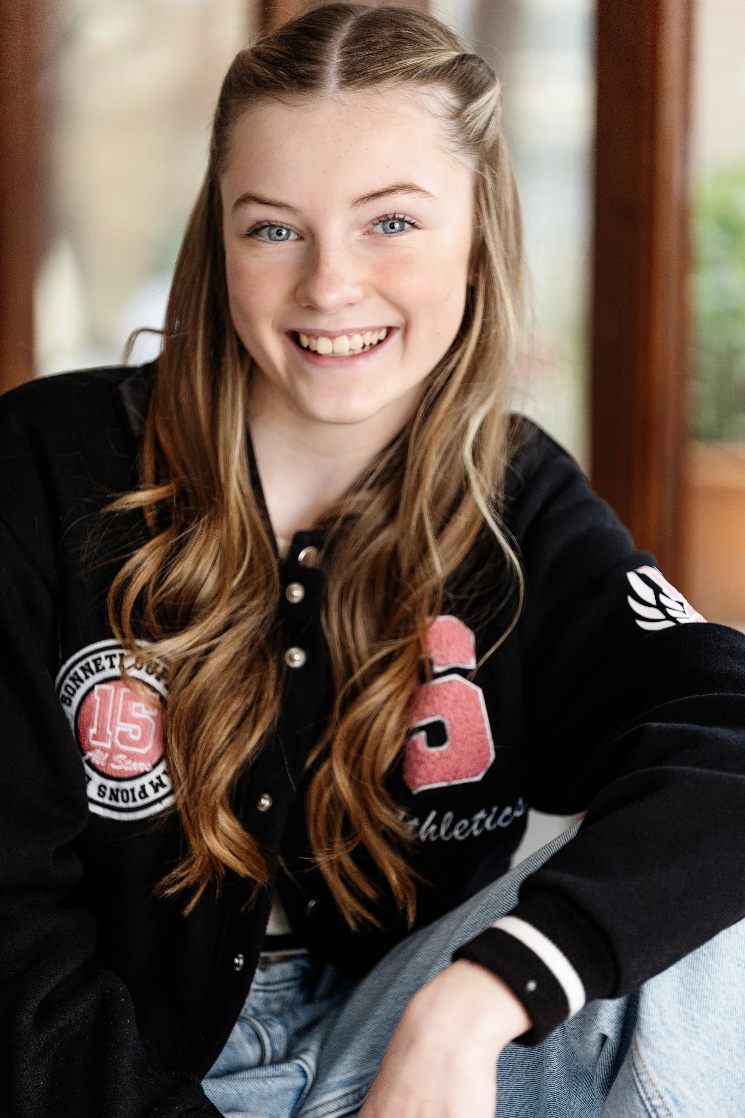 A young woman with long blonde hair wearing a black jacket with patches and lettering, smiling and looking at the camera indoors with wooden elements and windows in the background.