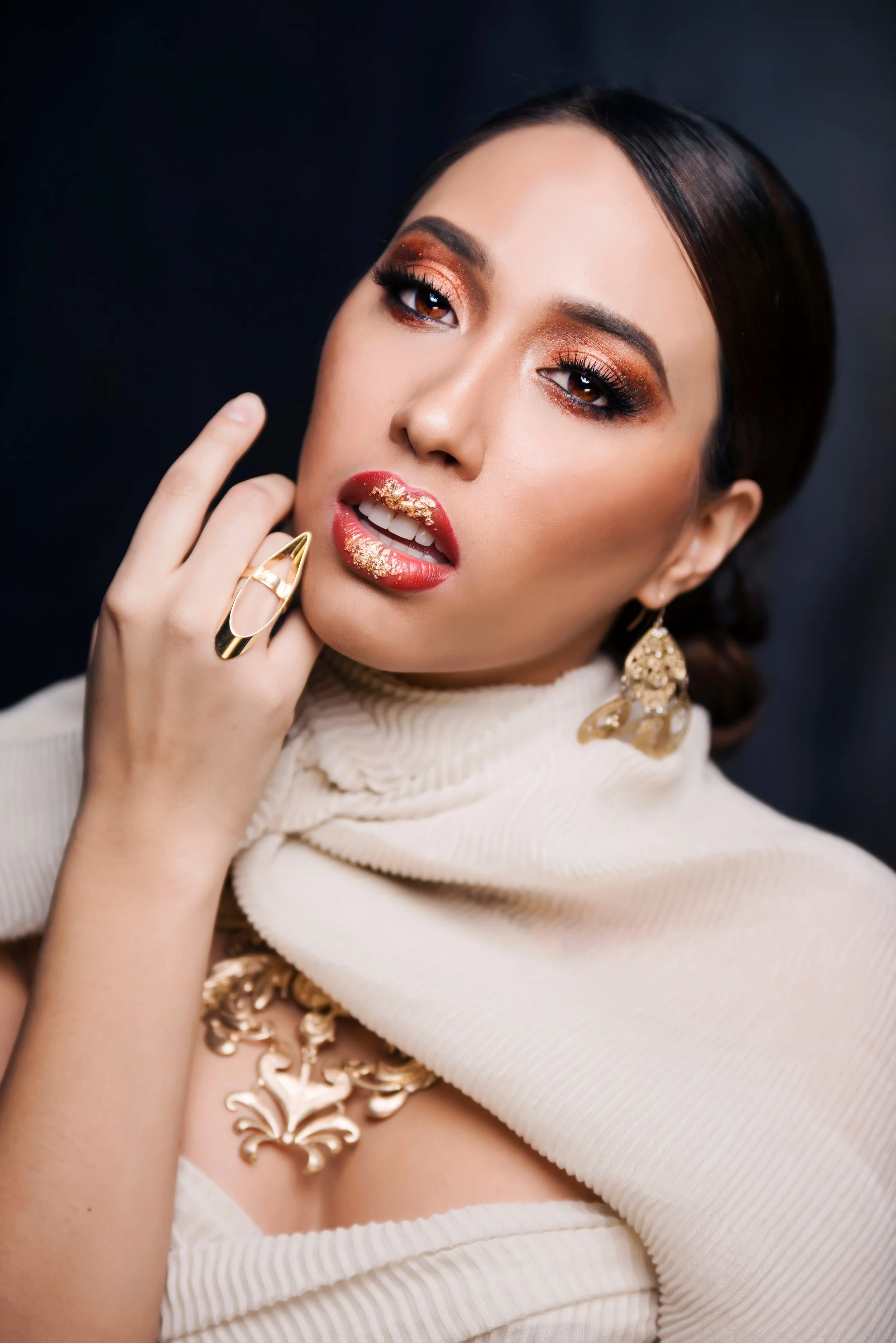 A woman with dark hair and brown eyes wearing gold earrings and a cream-colored sweater with gold decorative accents. She has on bronze eyeshadow, long dark eyelashes, and red lips with gold glitter. Her hand with a large gold ring touches her face.